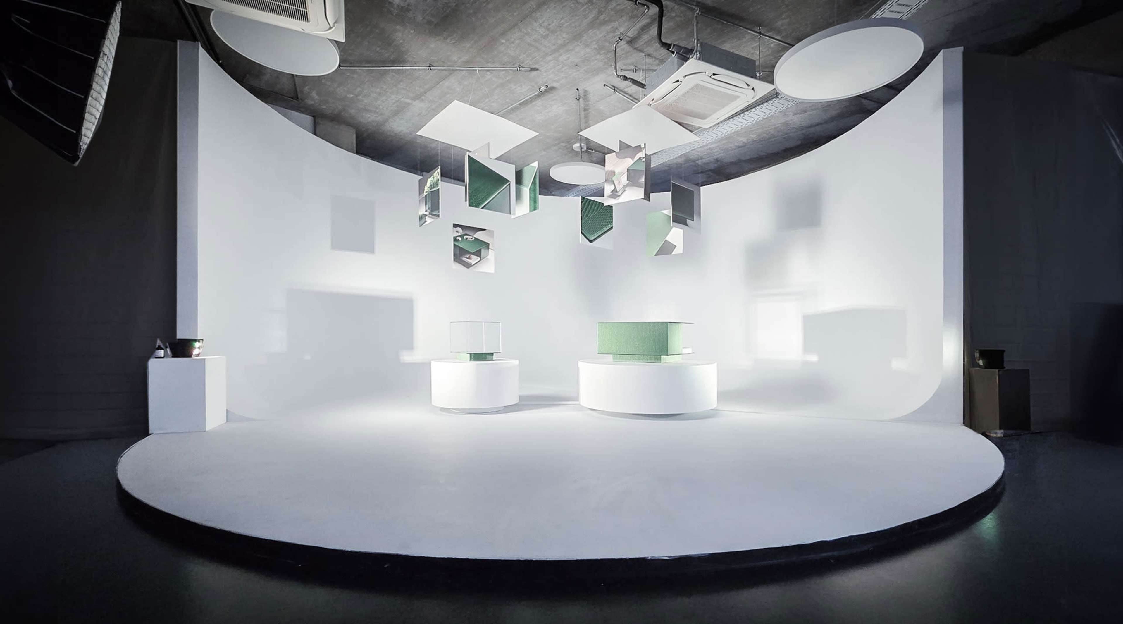 The image shows a minimalist exhibition space with circular platforms and geometric shapes hanging from the ceiling against a white backdrop.