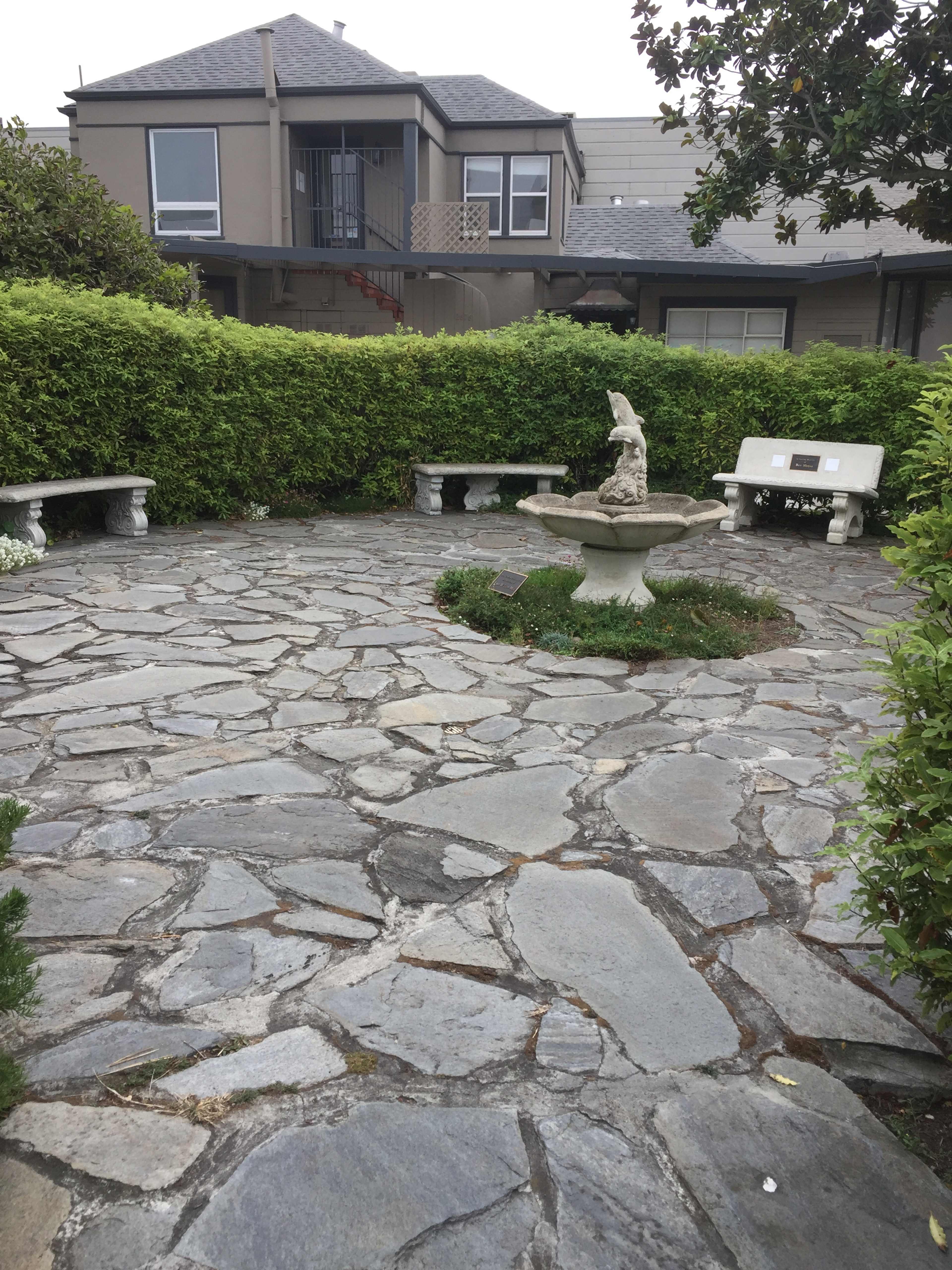 A stone-paved courtyard featuring a central fountain and surrounded by benches and trimmed hedges.