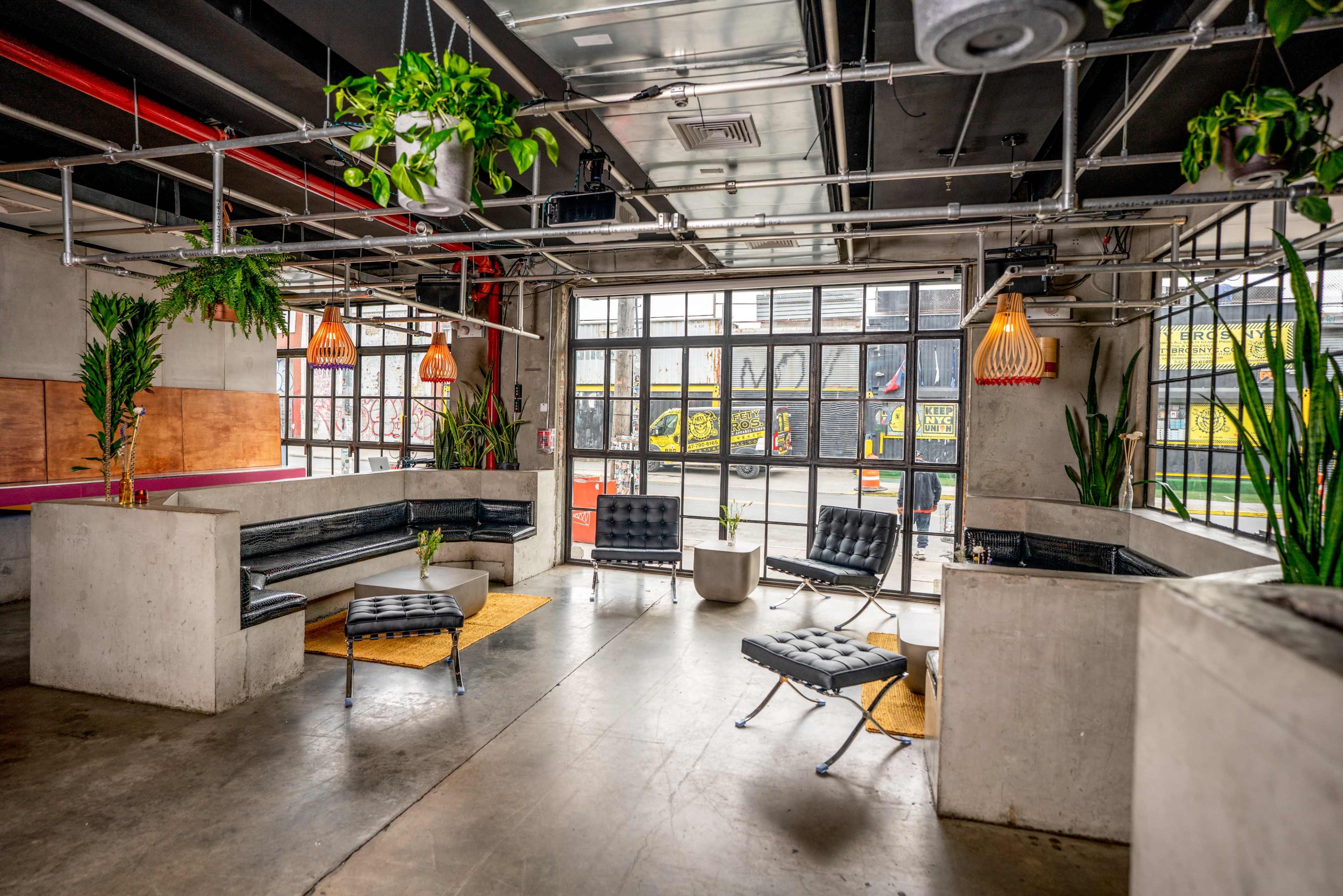 The image shows a modern lounge area with concrete walls, seating arrangements, and plants, featuring large windows that open up to an outdoor view.