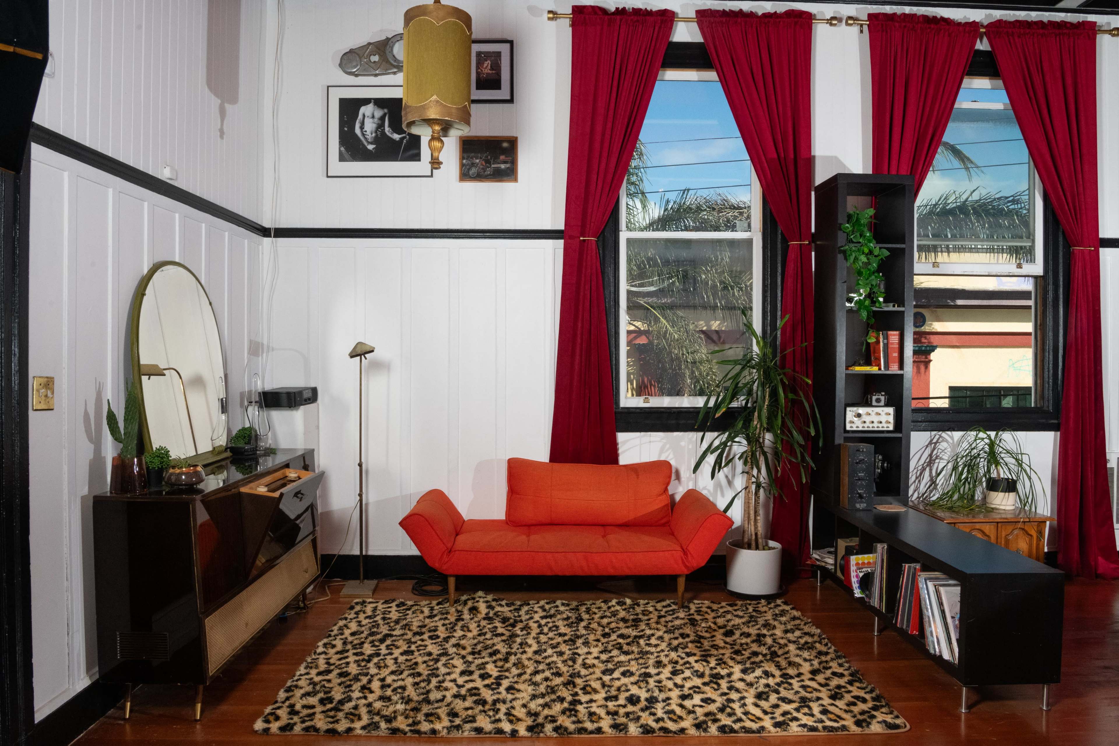 A living room with an orange couch, a mirror, a record shelf, and large windows framed by red curtains.