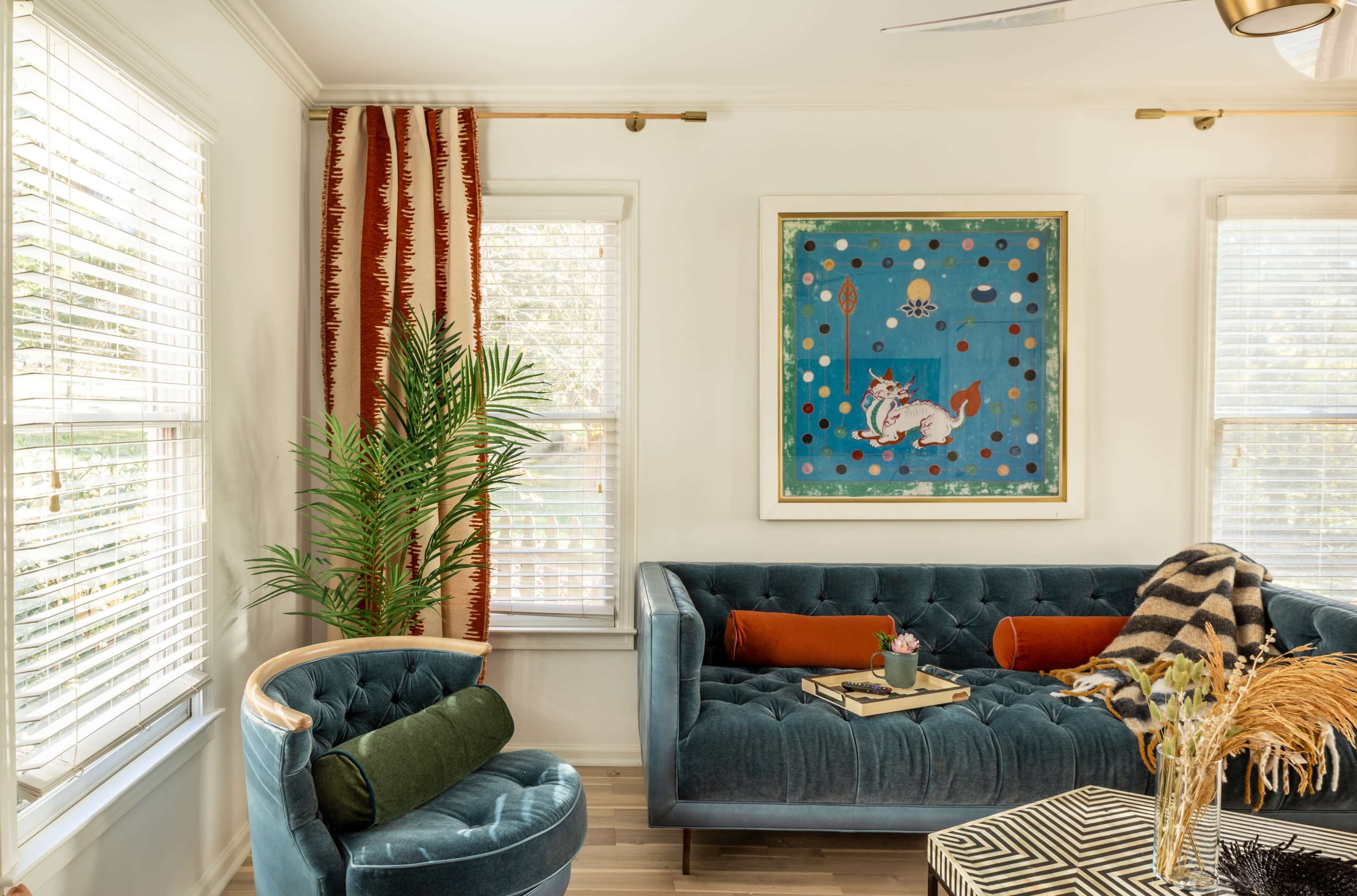 A bright living room features a blue velvet sofa, a round upholstered chair, and a colorful wall art piece amidst large windows with light-filtering curtains.
