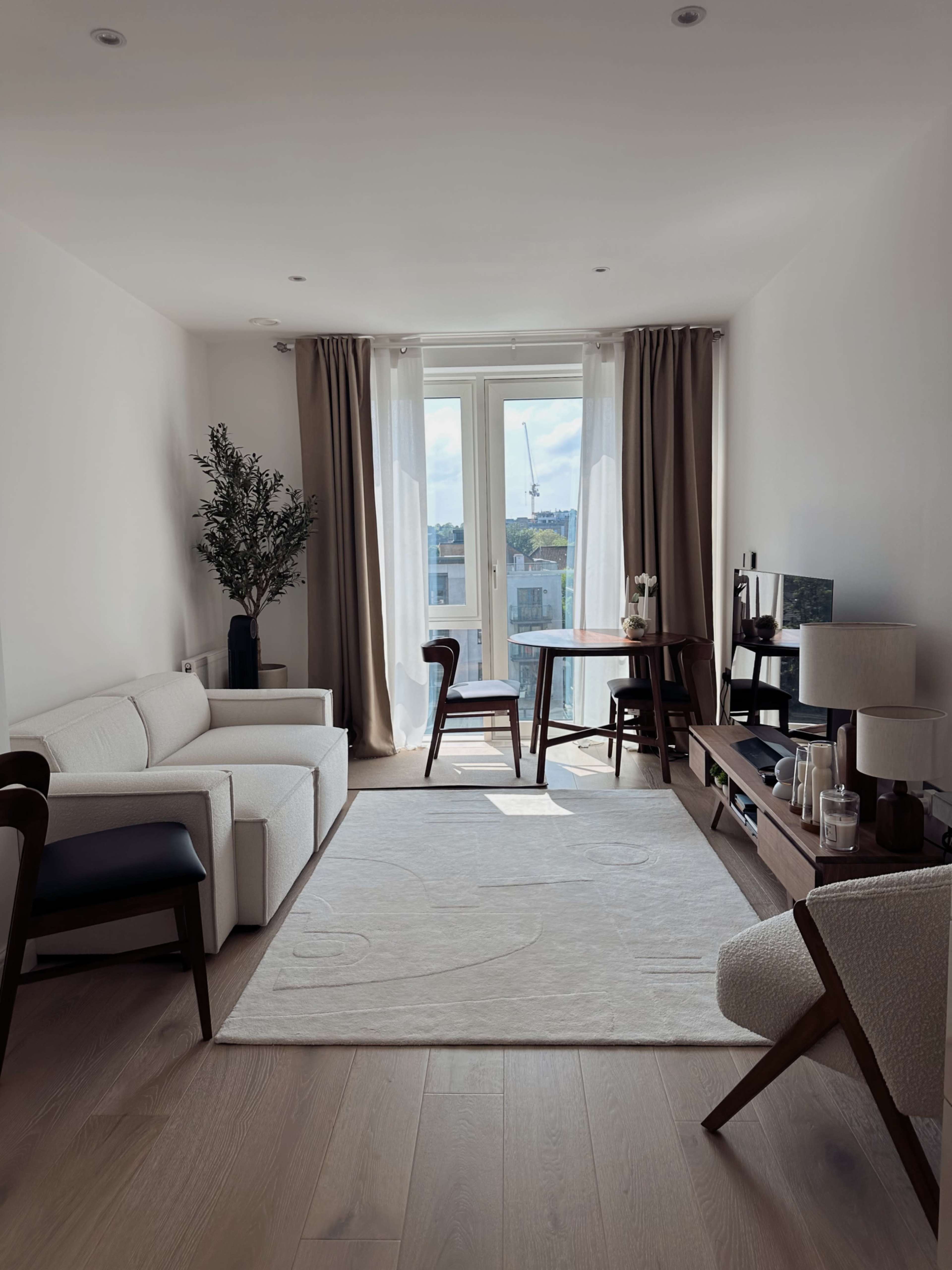 The image depicts a modern living room featuring a white sofa, two wooden chairs, a small round table, and large windows with curtains.