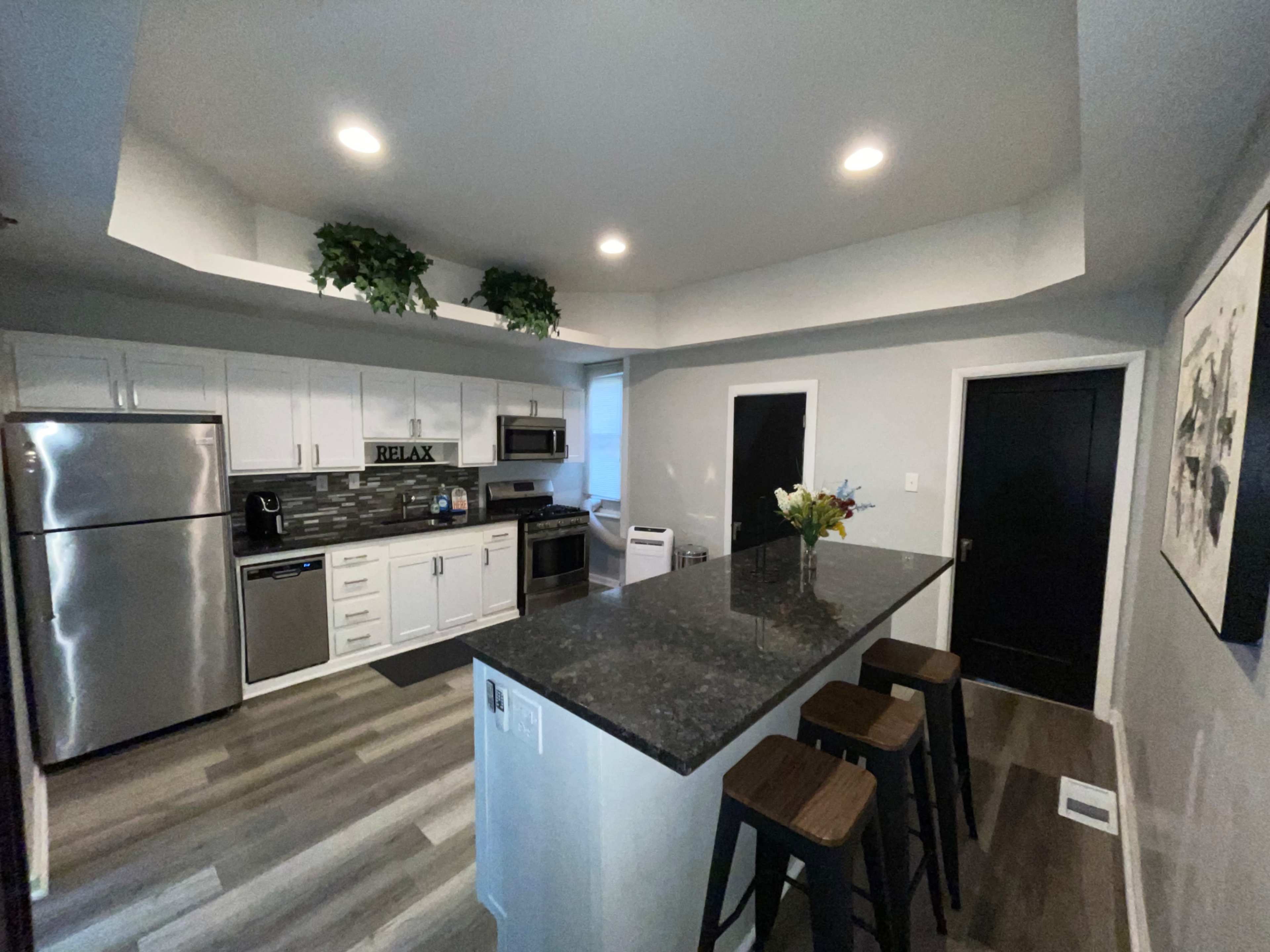 The image shows a modern kitchen with stainless steel appliances, white cabinetry, and a dark countertop with bar stools at a raised island.