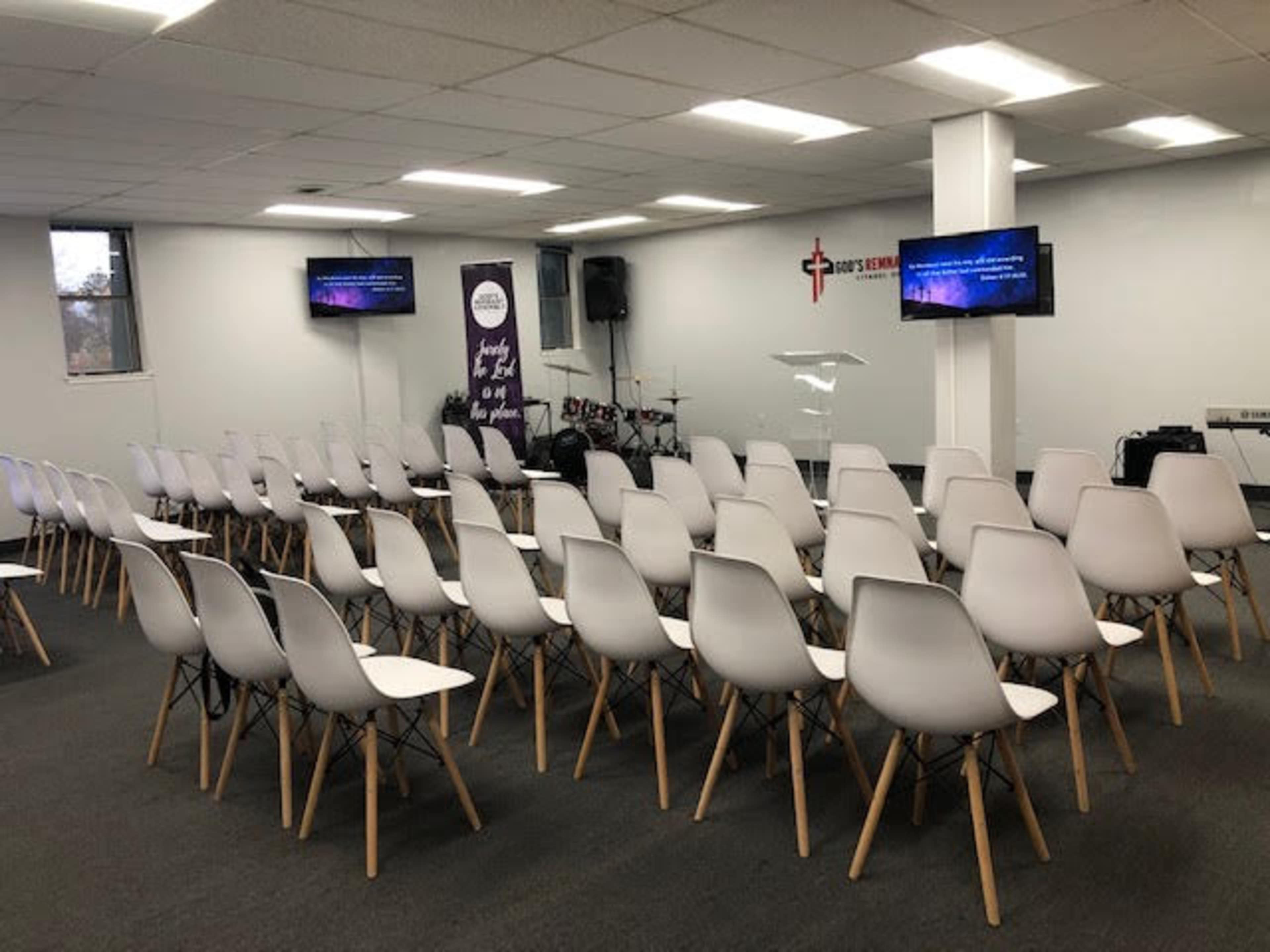 An empty indoor space set up with rows of white seating facing a clear podium and two screens displaying text, with musical instruments visible in the background.