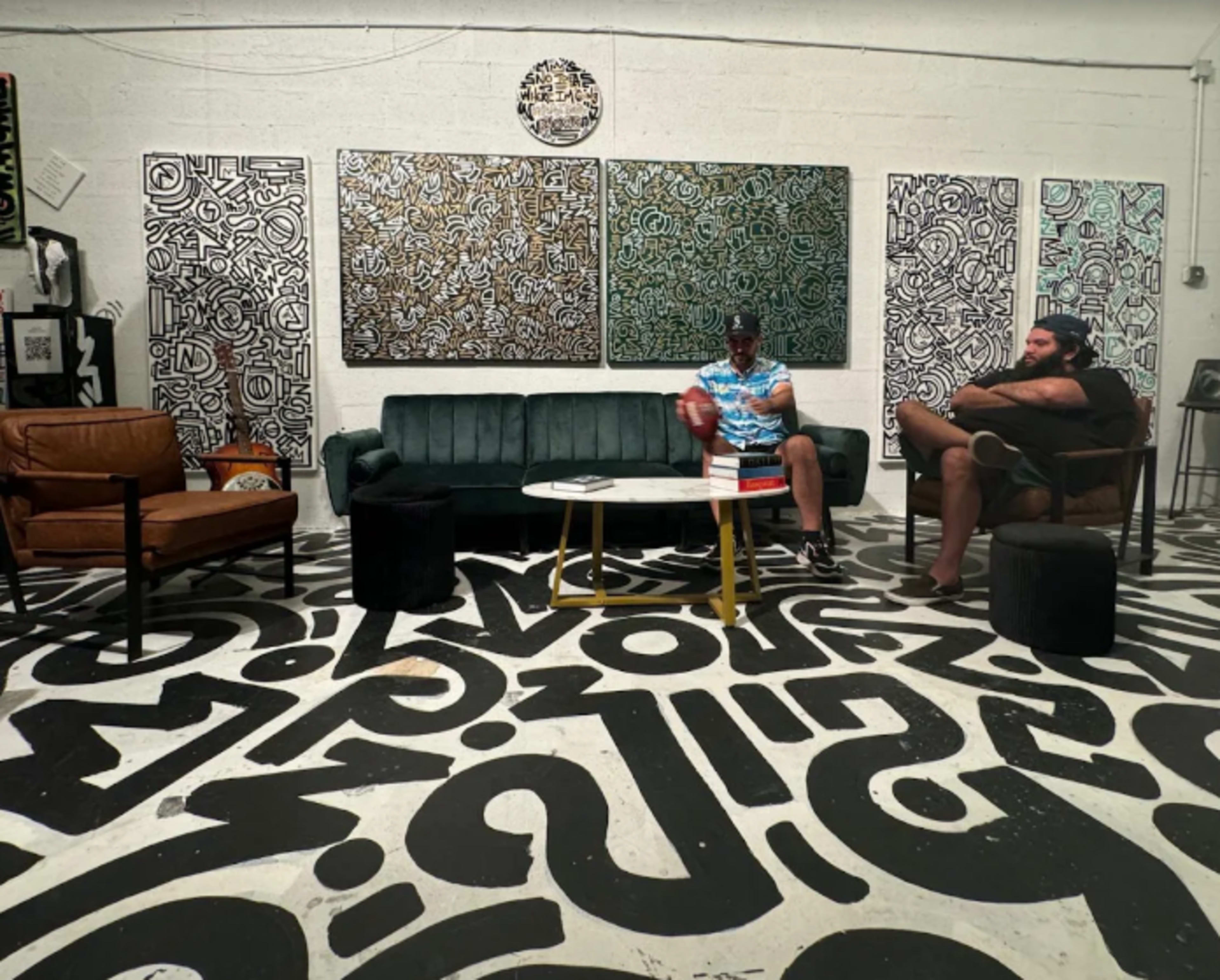 Two men are seated in a modern art gallery with bold black and white patterned flooring and colorful abstract artworks on the walls.