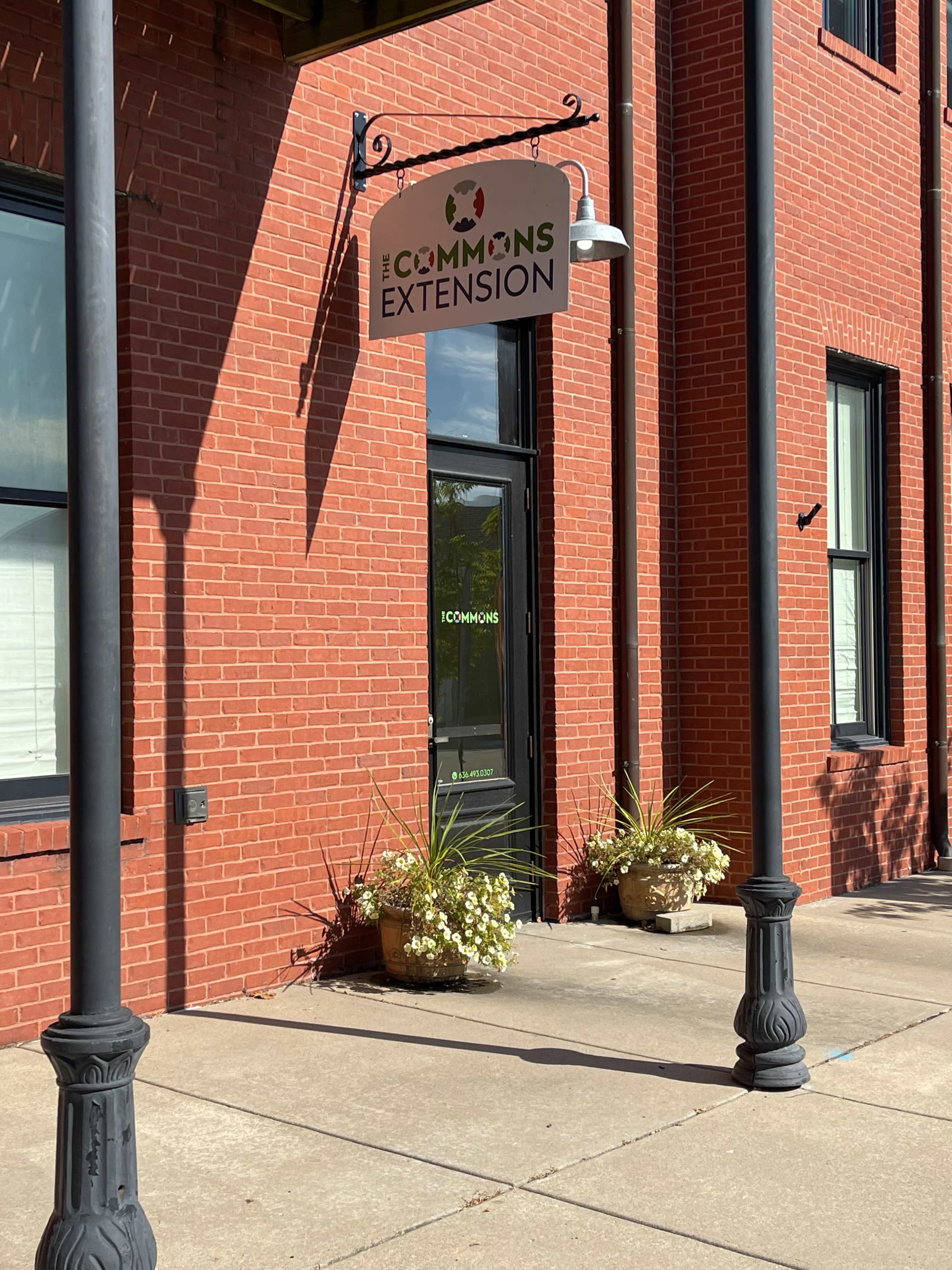 The image shows a brick building entrance with a sign reading "The Commons Extension" and flower pots on either side of the door.