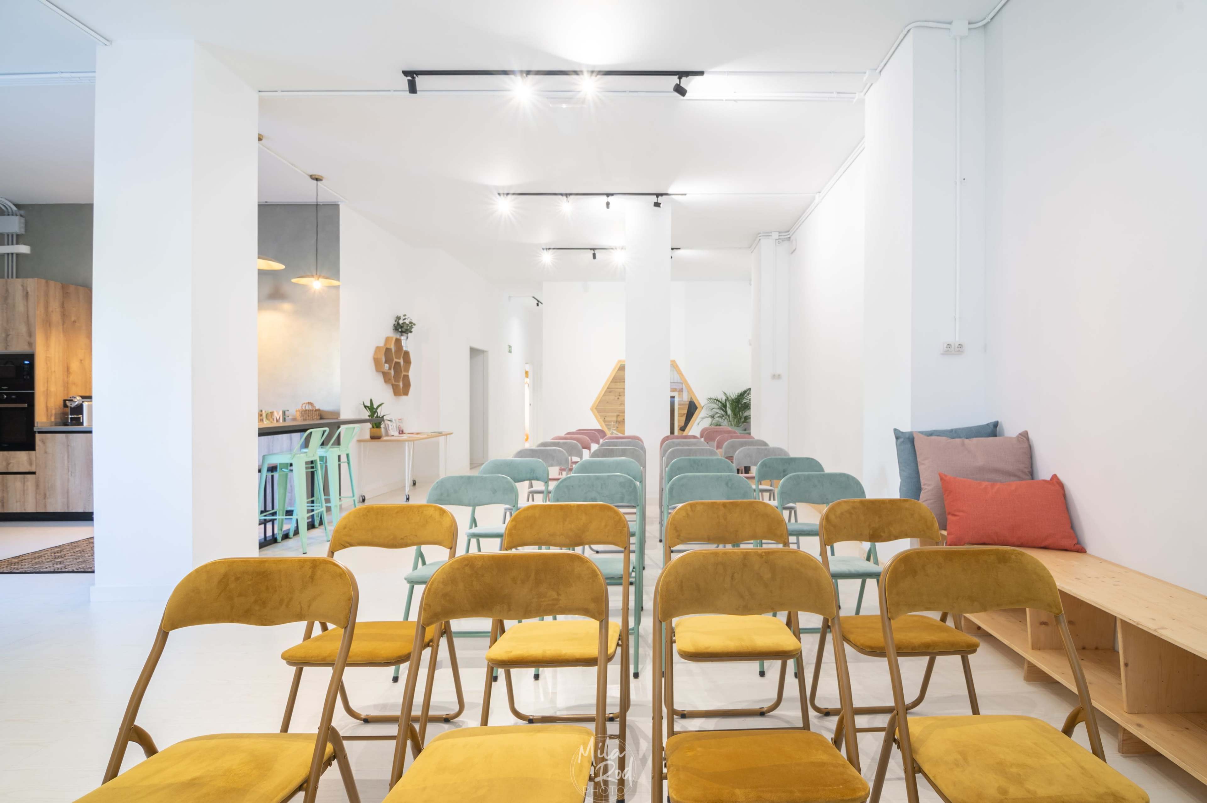 A row of colorful folding chairs arranged facing a minimalist design space with a kitchen area in the background.