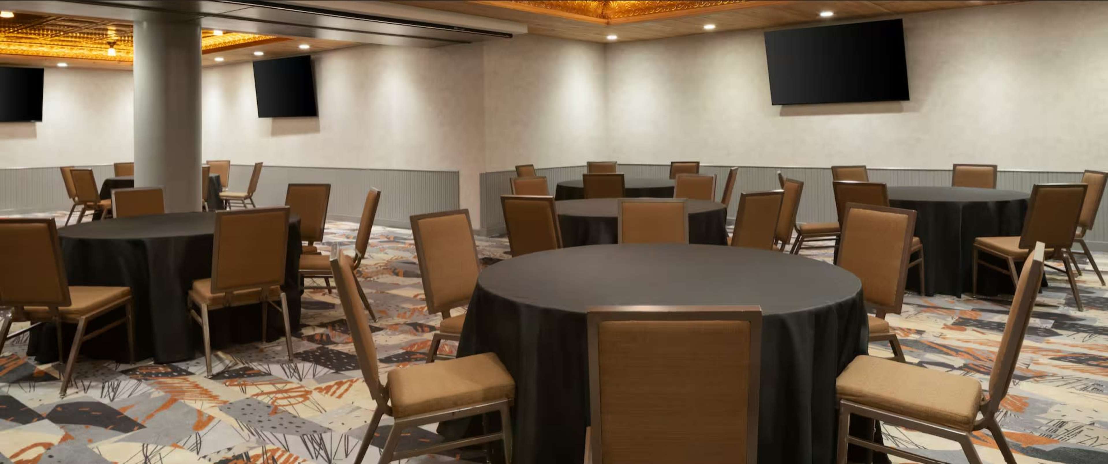 The image shows a conference room with several round tables and chairs arranged neatly on a patterned carpet, with two screens mounted on the walls.