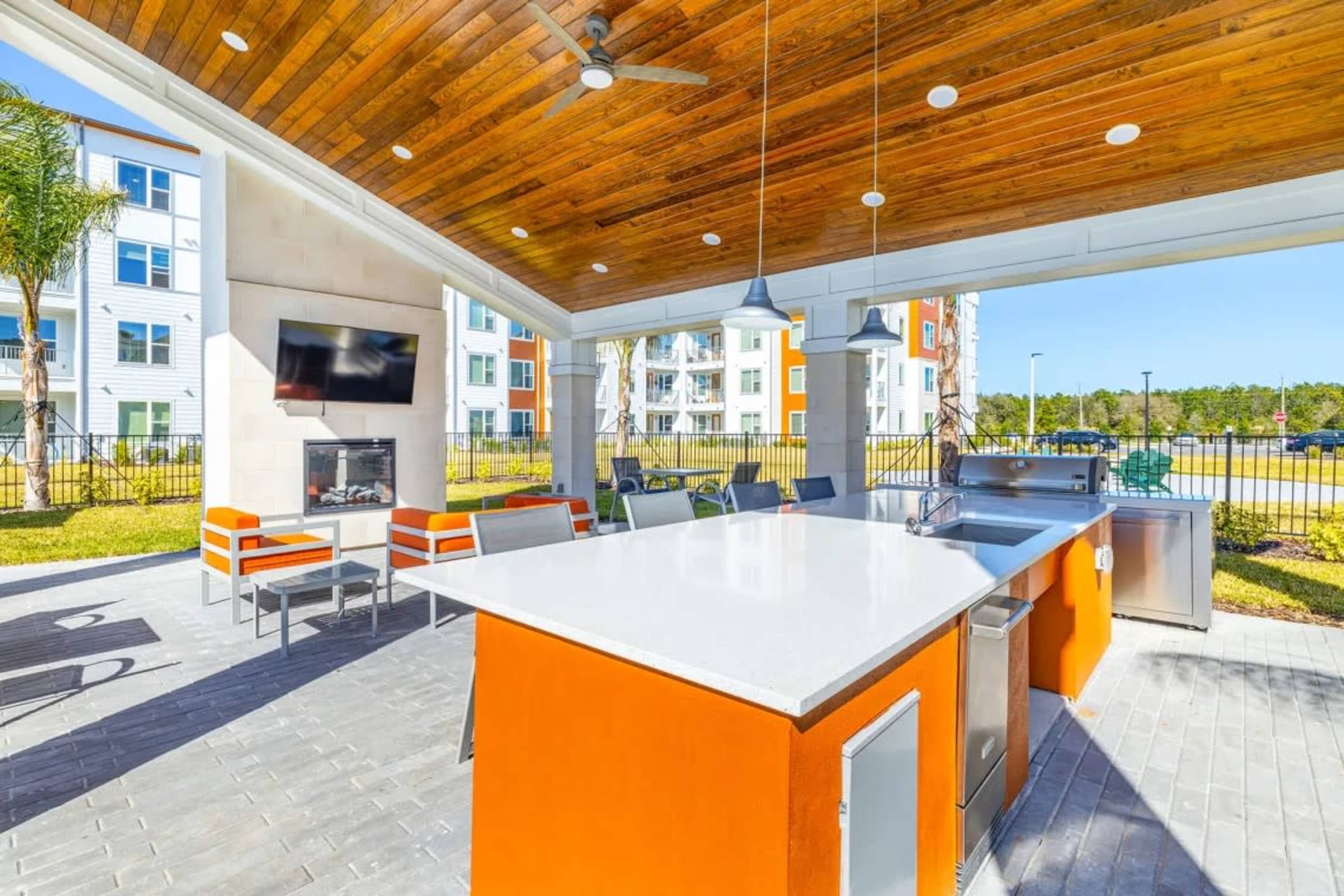 The image shows a modern outdoor kitchen area with a large countertop, grill, and seating, under a wooden roof with a ceiling fan.