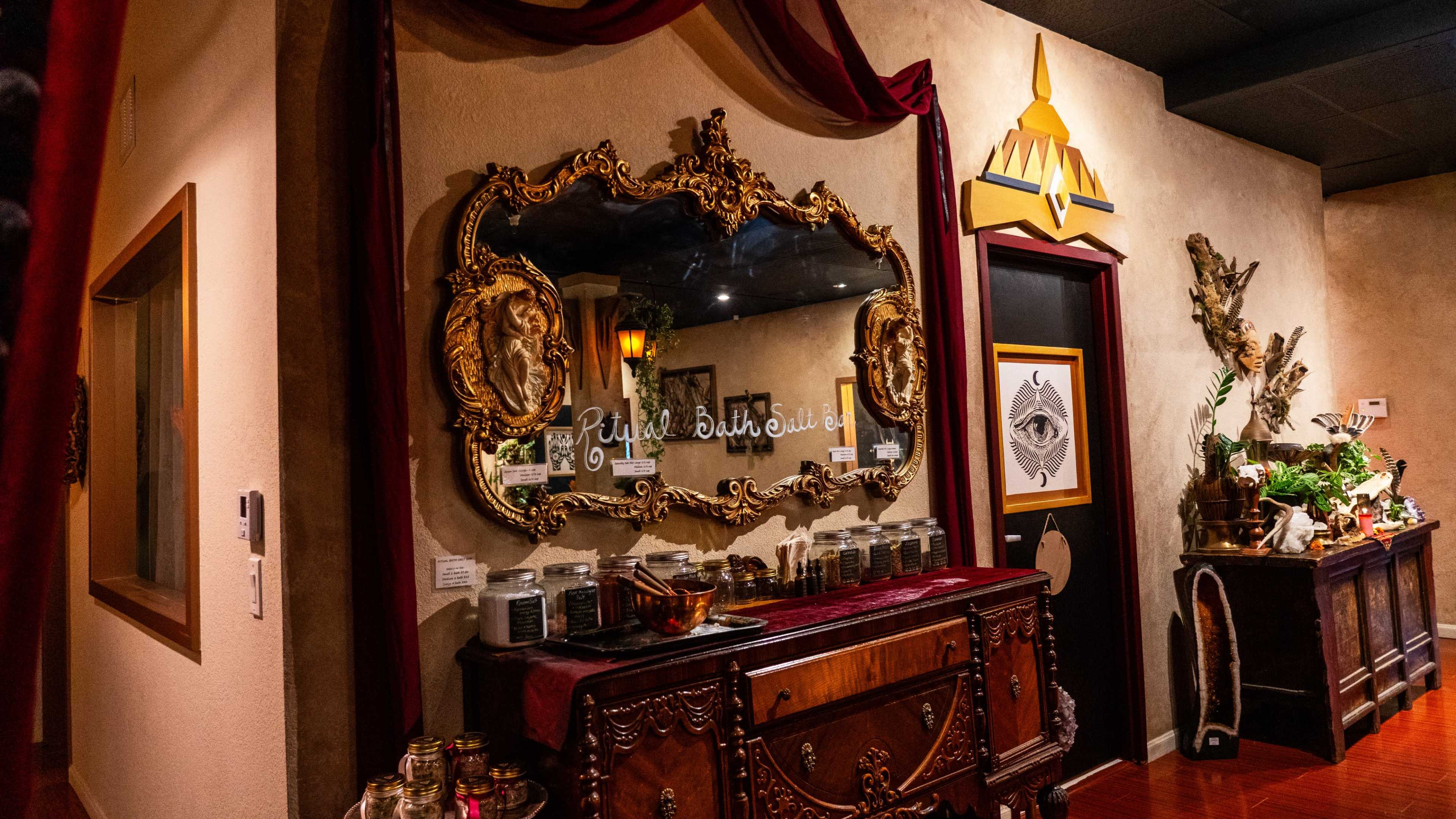 The image shows a warmly lit room featuring an ornate mirror above a wooden dresser with various jars, alongside a door decorated with an intricate design.