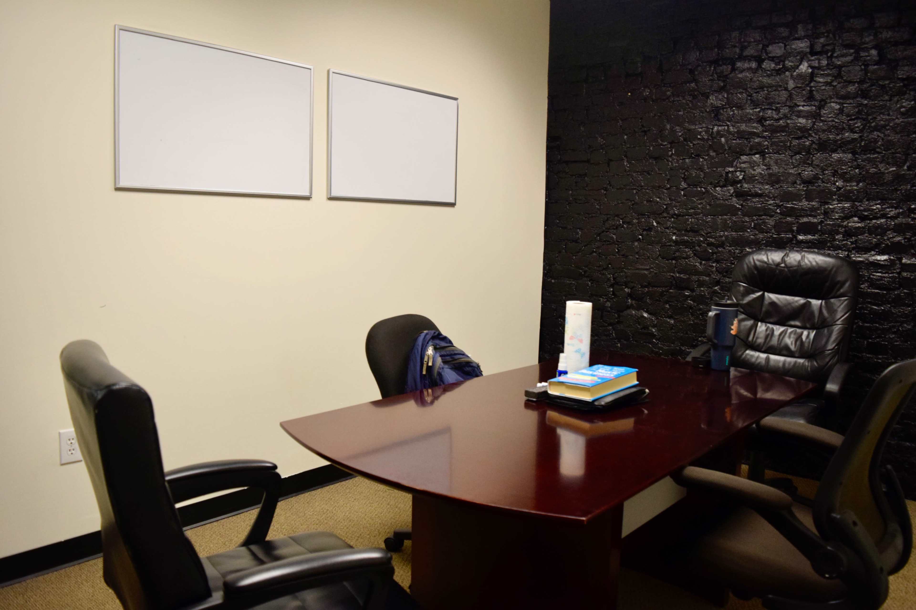 The image shows a small office with a dark brick wall, a wooden table, two office chairs, and two blank whiteboards mounted on the wall.