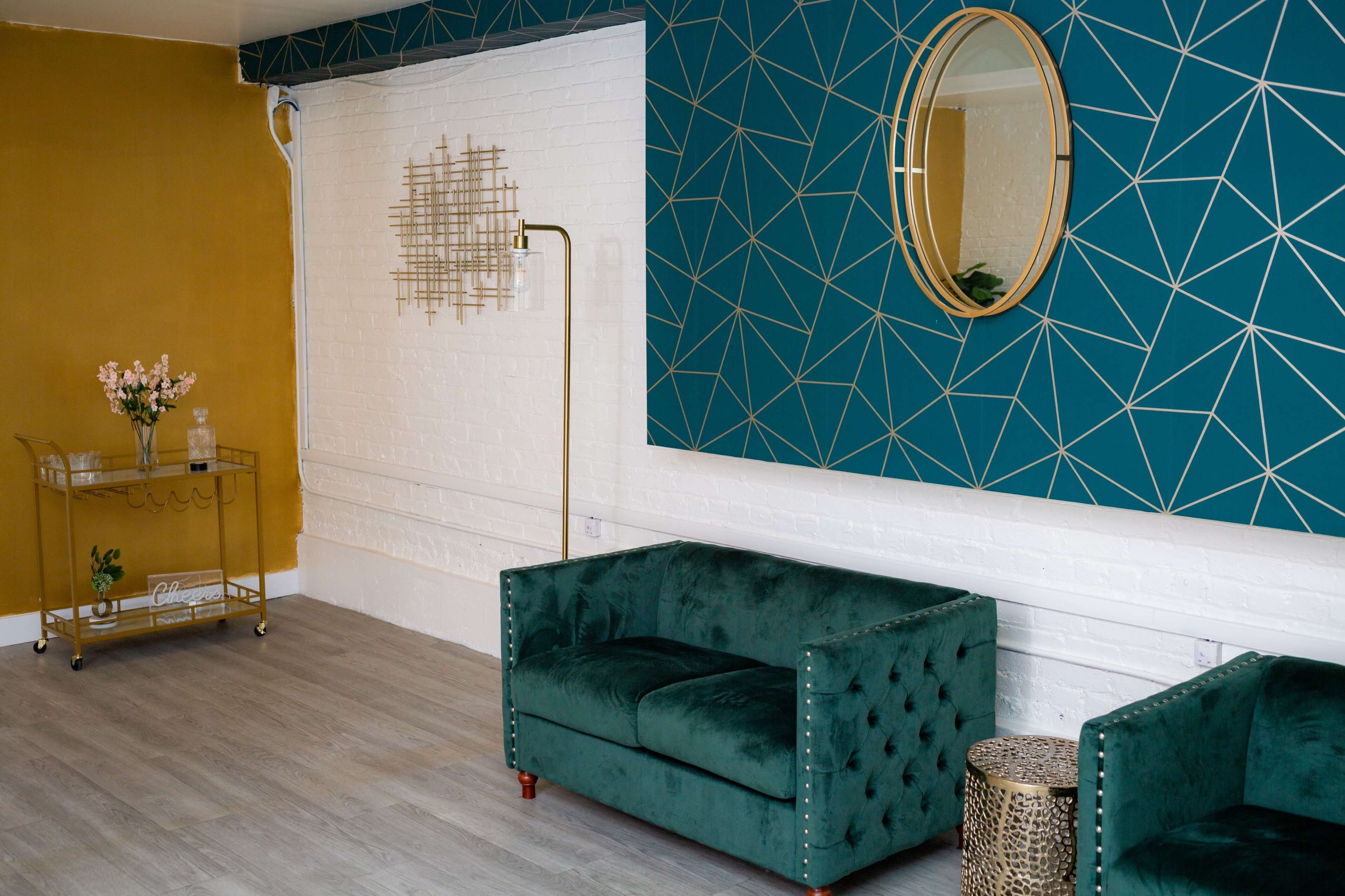 The image shows a modern interior with a teal geometric accent wall, a golden side table with flowers, and a green upholstered sofa.
