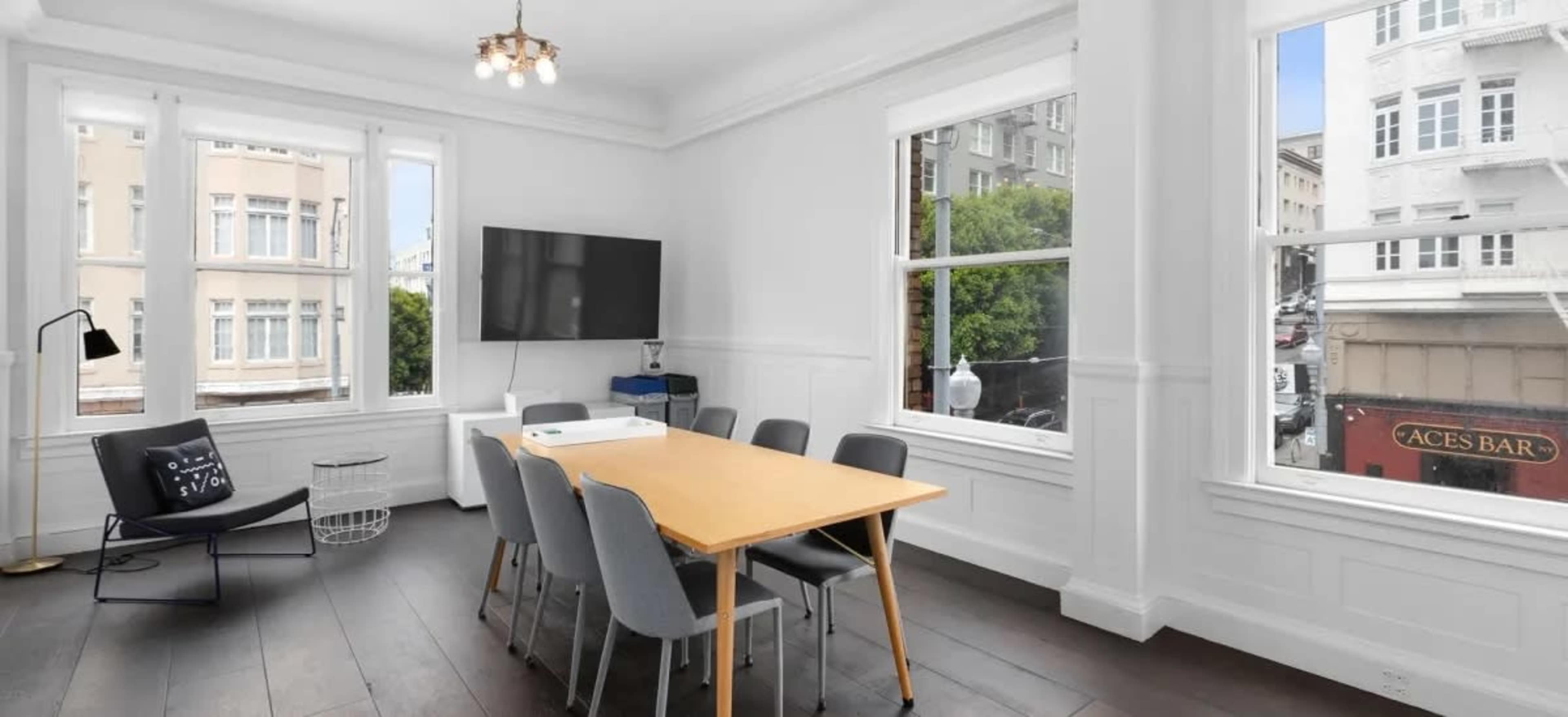 A bright, minimalist dining area featuring a light wooden table surrounded by gray chairs, with large windows offering a view of the street below.