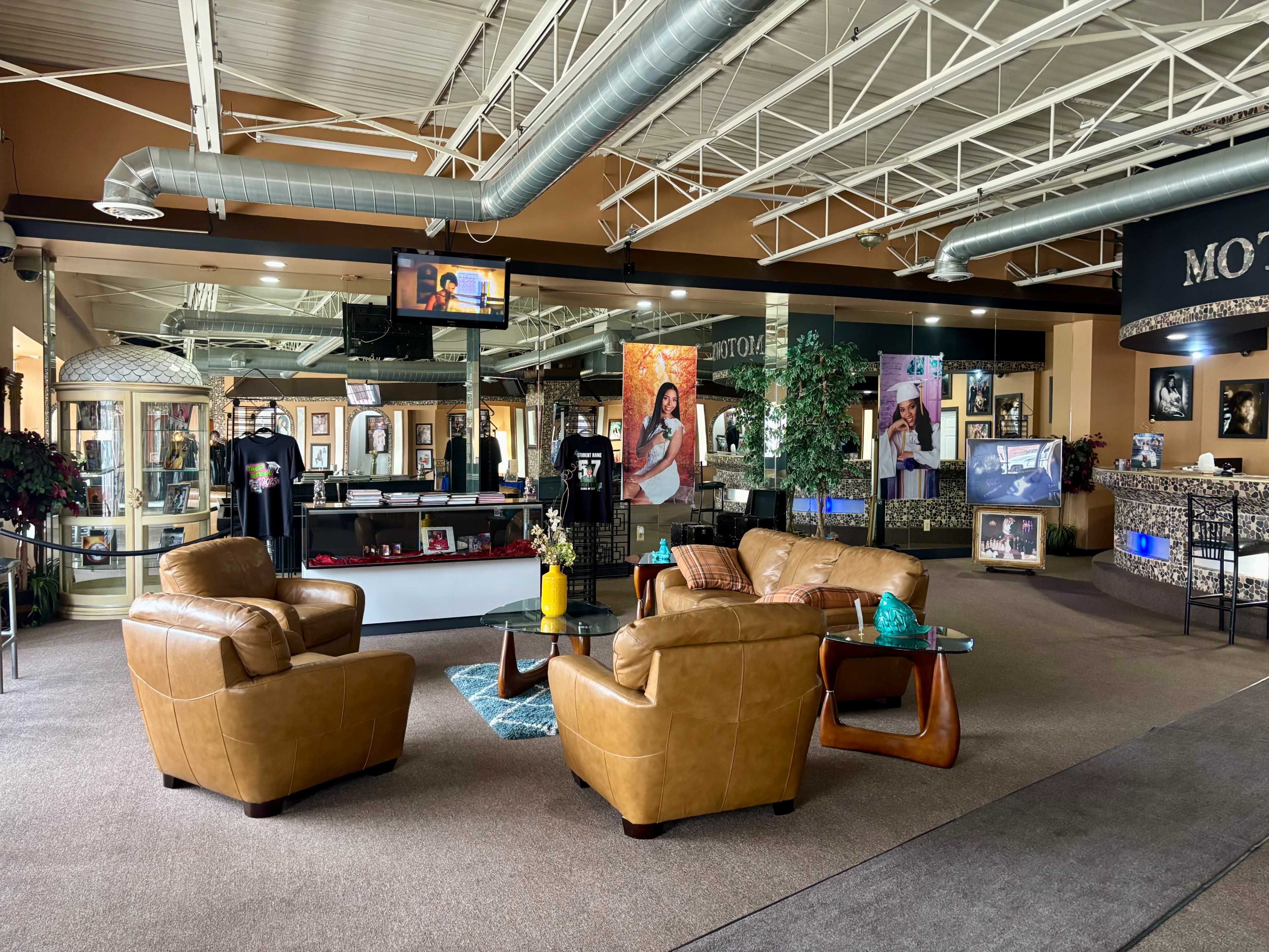 The image shows a spacious, well-lit interior of a facility featuring a seating area with tan leather chairs and a decorative table, surrounded by various displays and photographs on the walls.