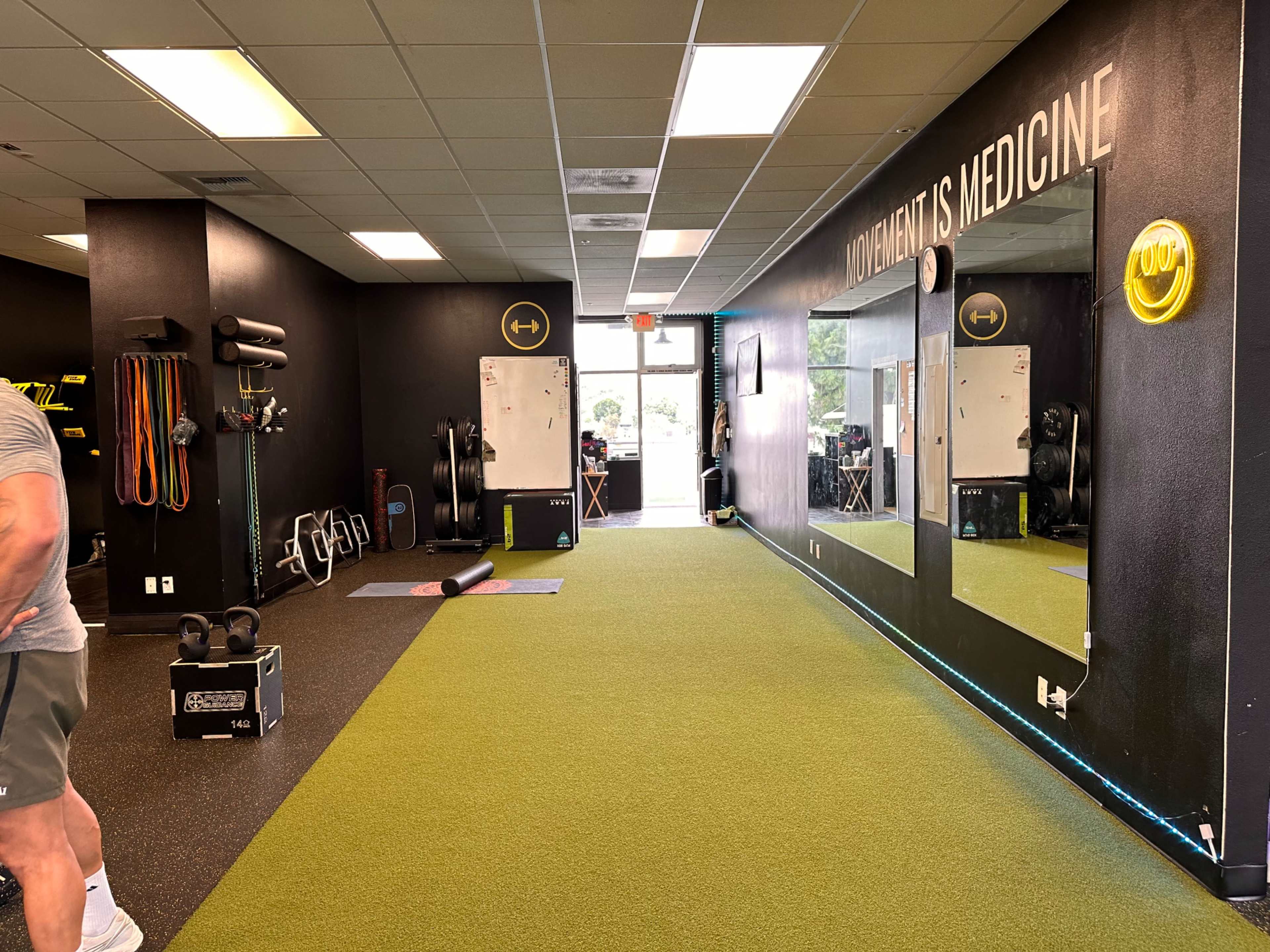 A fitness gym interior with green turf flooring, equipment on the walls, and large mirrors reflecting a doorway leading outside.