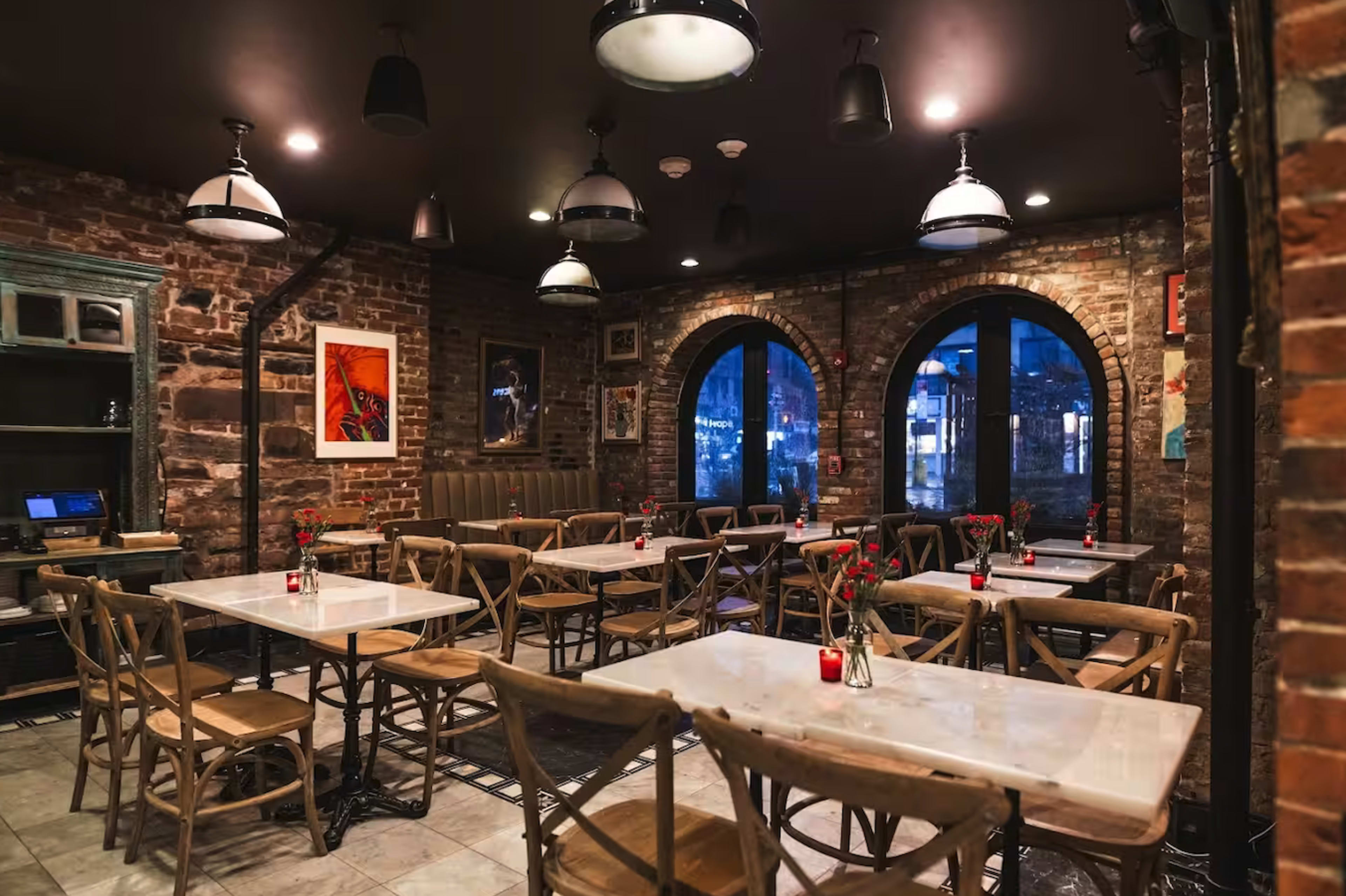 The image shows a brick-walled restaurant interior with several wooden tables and chairs, dim lighting, and large arched windows.