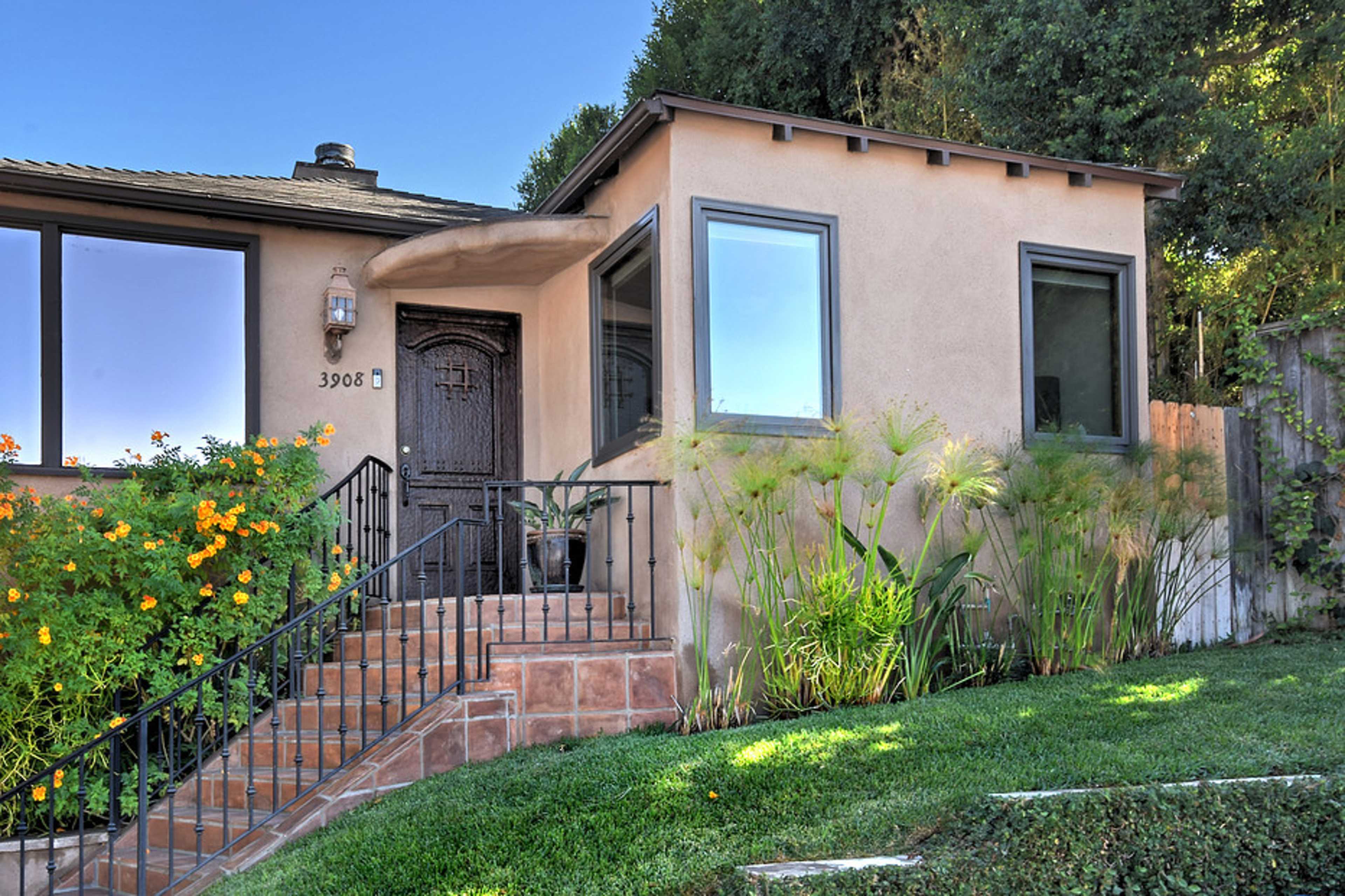 A single-story house with a decorative front door, surrounded by greenery and flowers, features a set of stairs leading up to the entryway.