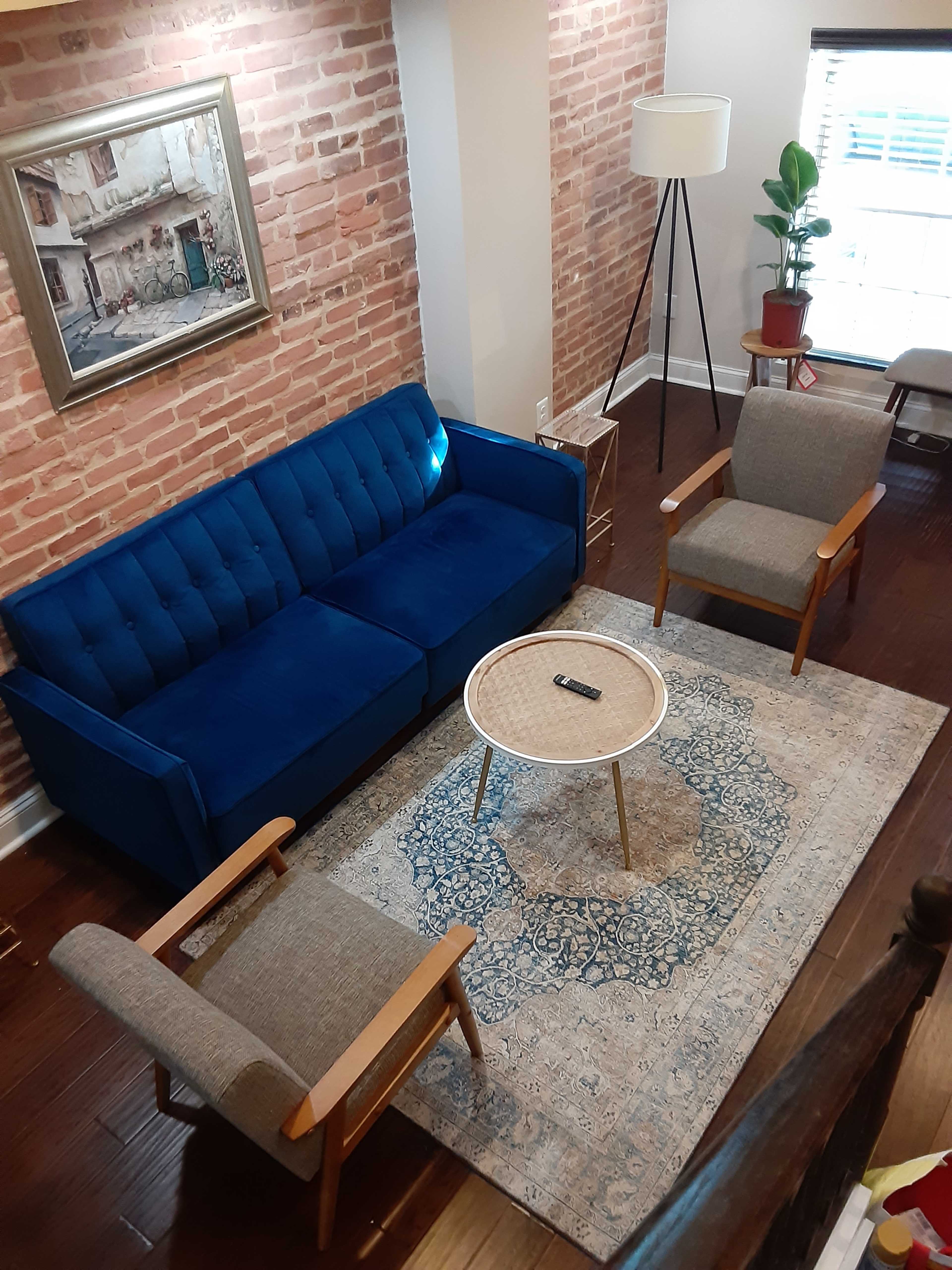 A living room with a blue sofa, two gray armchairs, a round coffee table, and a patterned rug, all set against a backdrop of exposed brick walls.