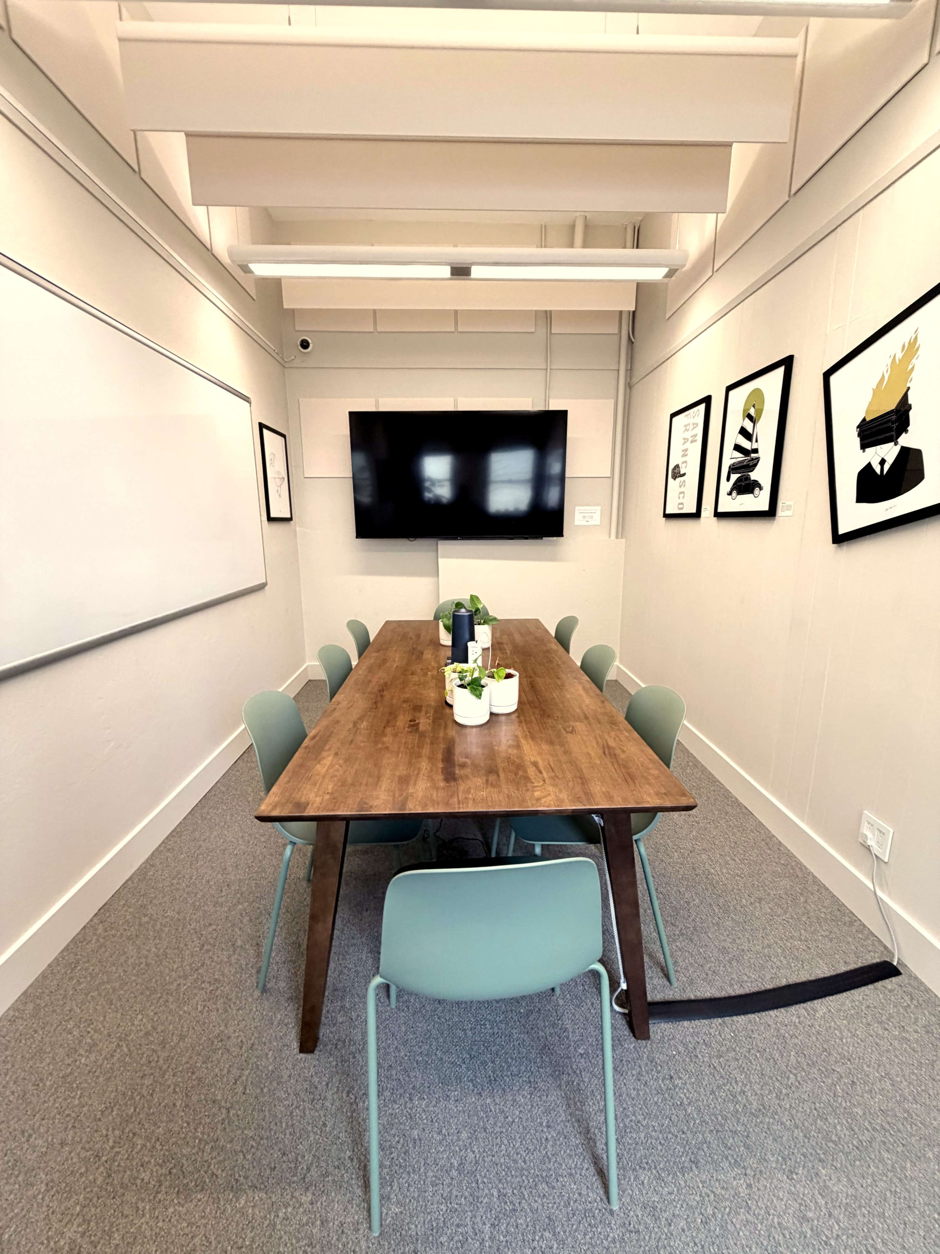 A conference room features a long wooden table surrounded by green chairs, with a TV mounted on the wall and framed art hanging nearby.