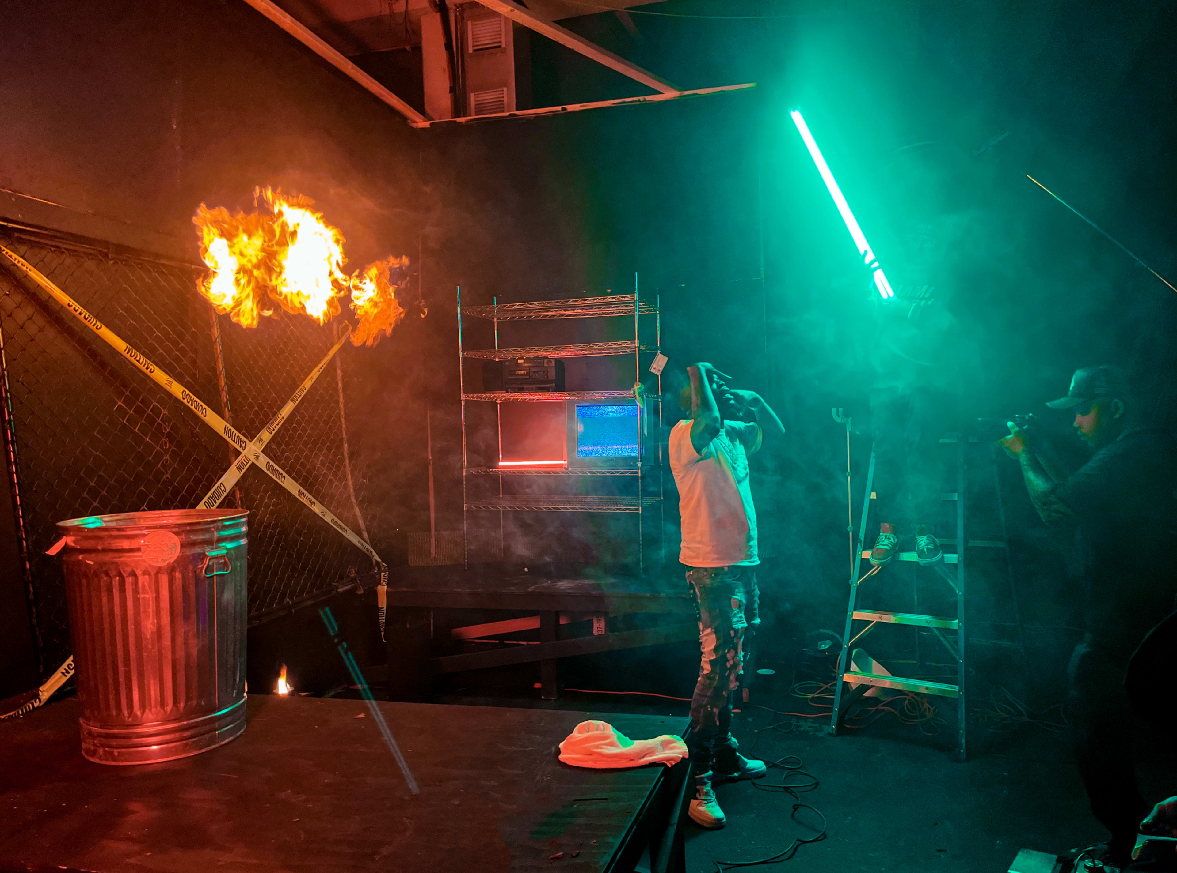 A person stands in a dimly lit room with smoke and colorful lighting, next to a flaming trash can and a camera setup on a ladder.