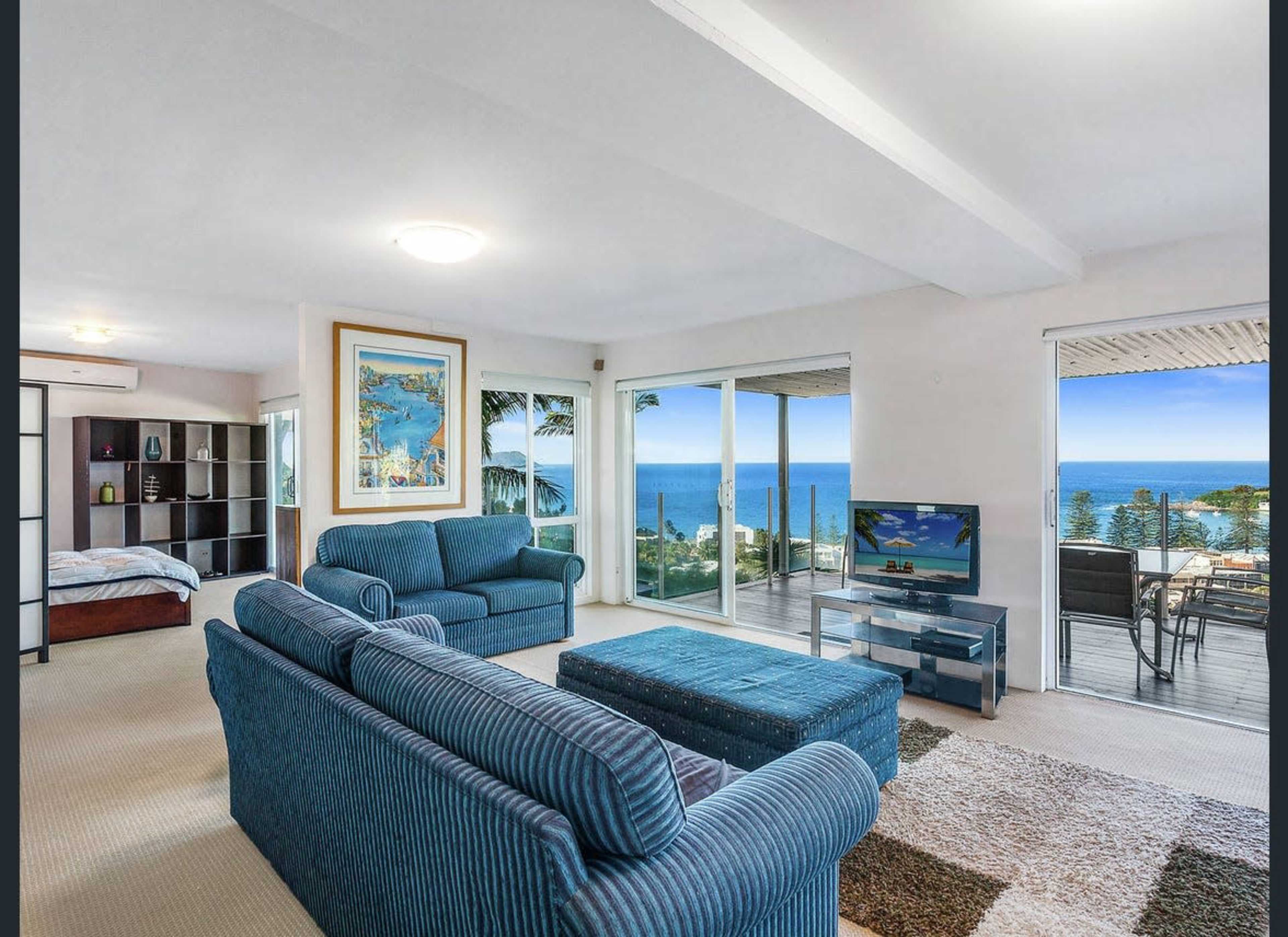 A spacious living room with two blue sofas, a television, and large windows overlooking a coastal view.