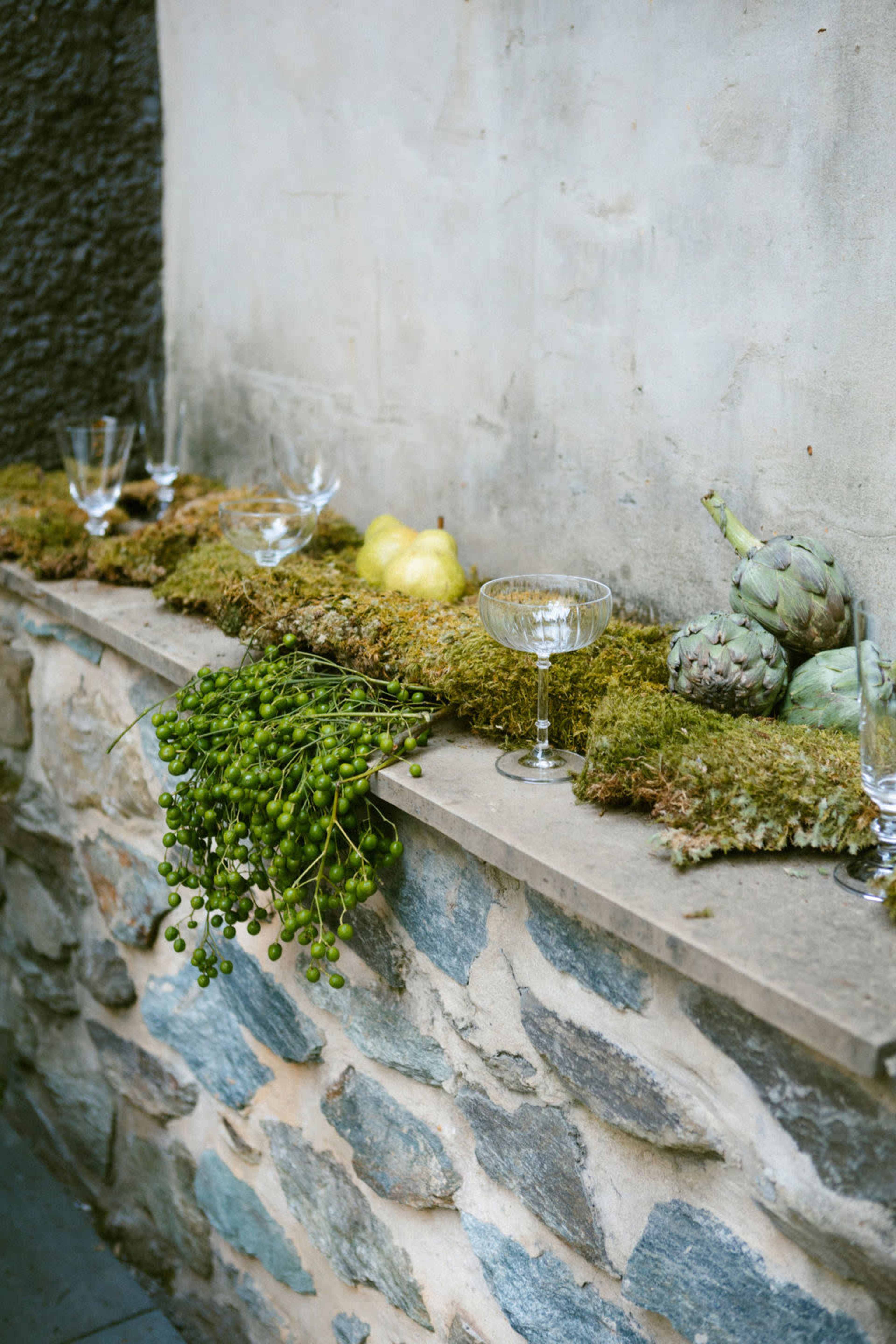 The image shows a stone ledge covered in moss, adorned with glassware, green grapes, and various decorative gourds.