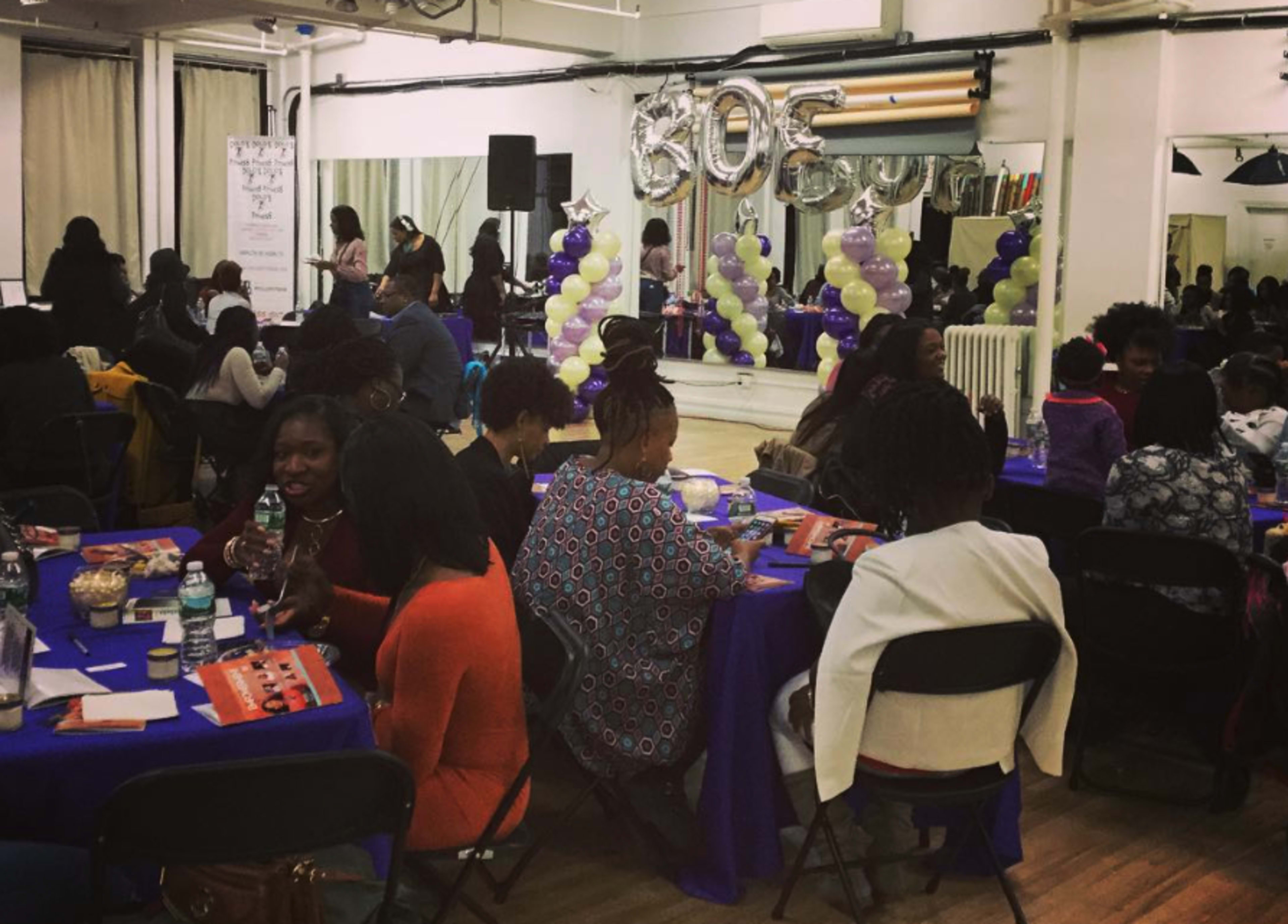 The image shows a gathering of people seated at tables with purple and gold decorations, celebrating an event in a spacious hall.
