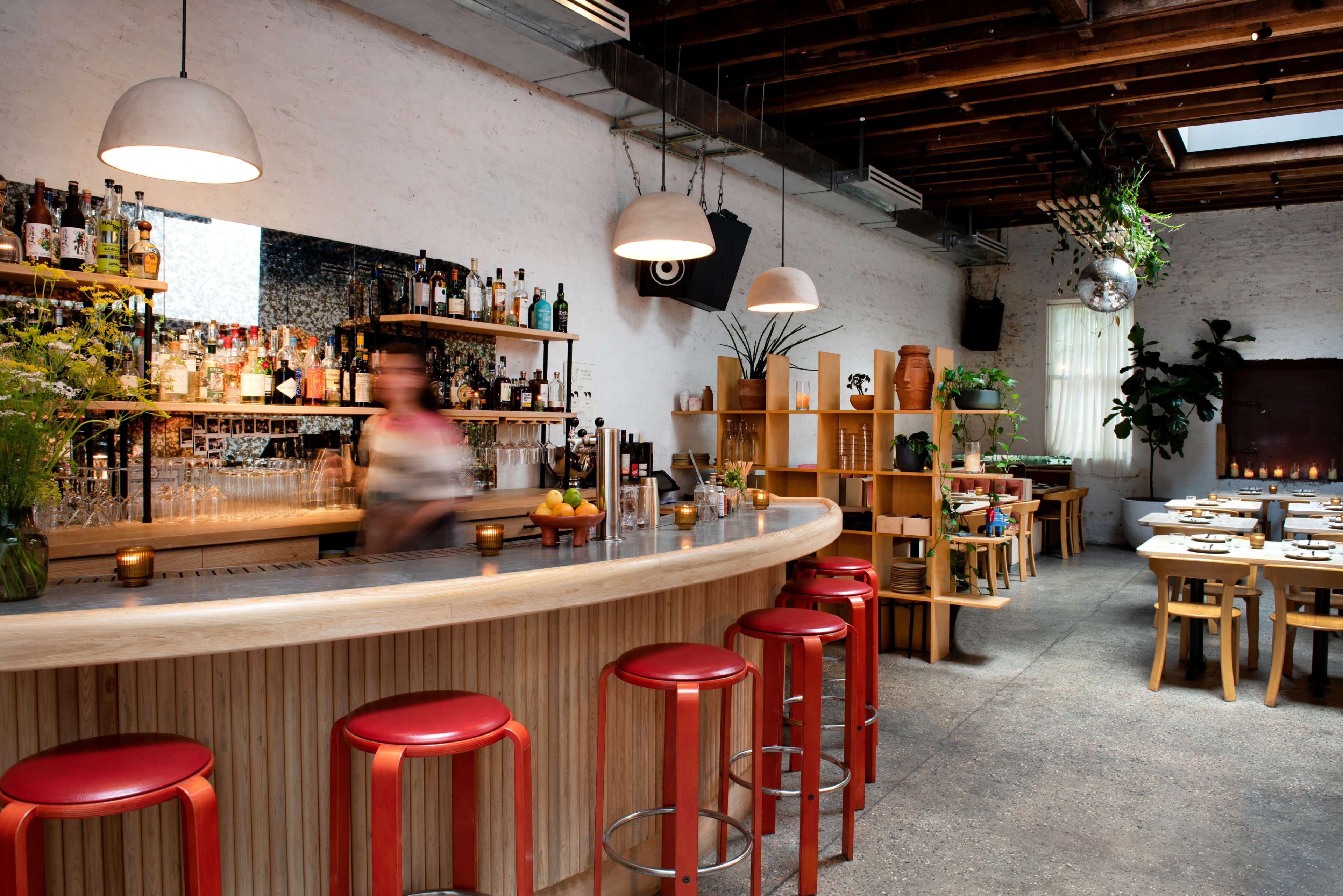 A modern bar area with a wooden countertop, red stools, and shelves filled with various bottles in a well-lit, minimalistic restaurant interior.