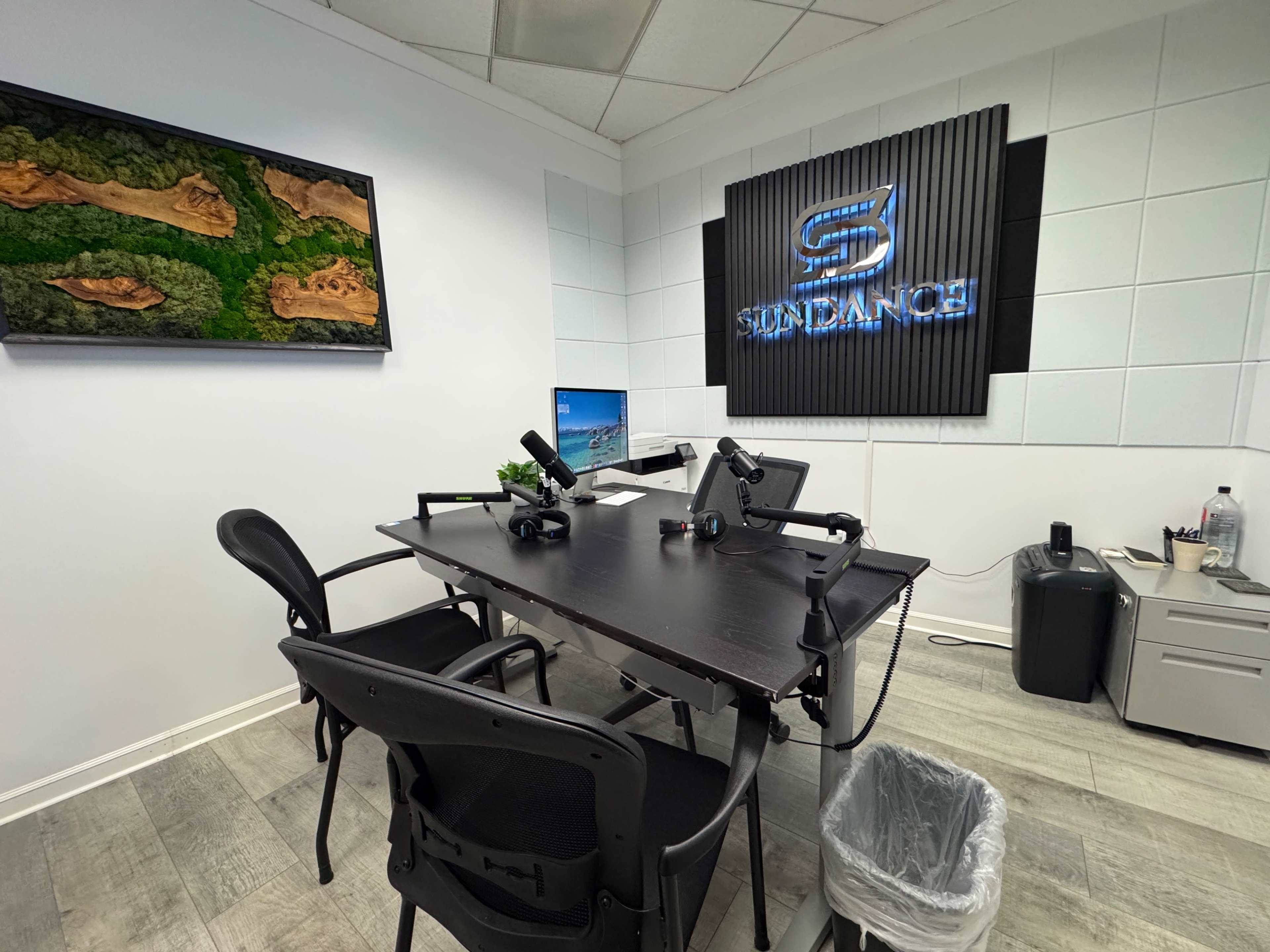 The image shows a modern office space featuring a black desk with chairs, a computer setup, a wall-mounted screen displaying a landscape, and a decorative sign.