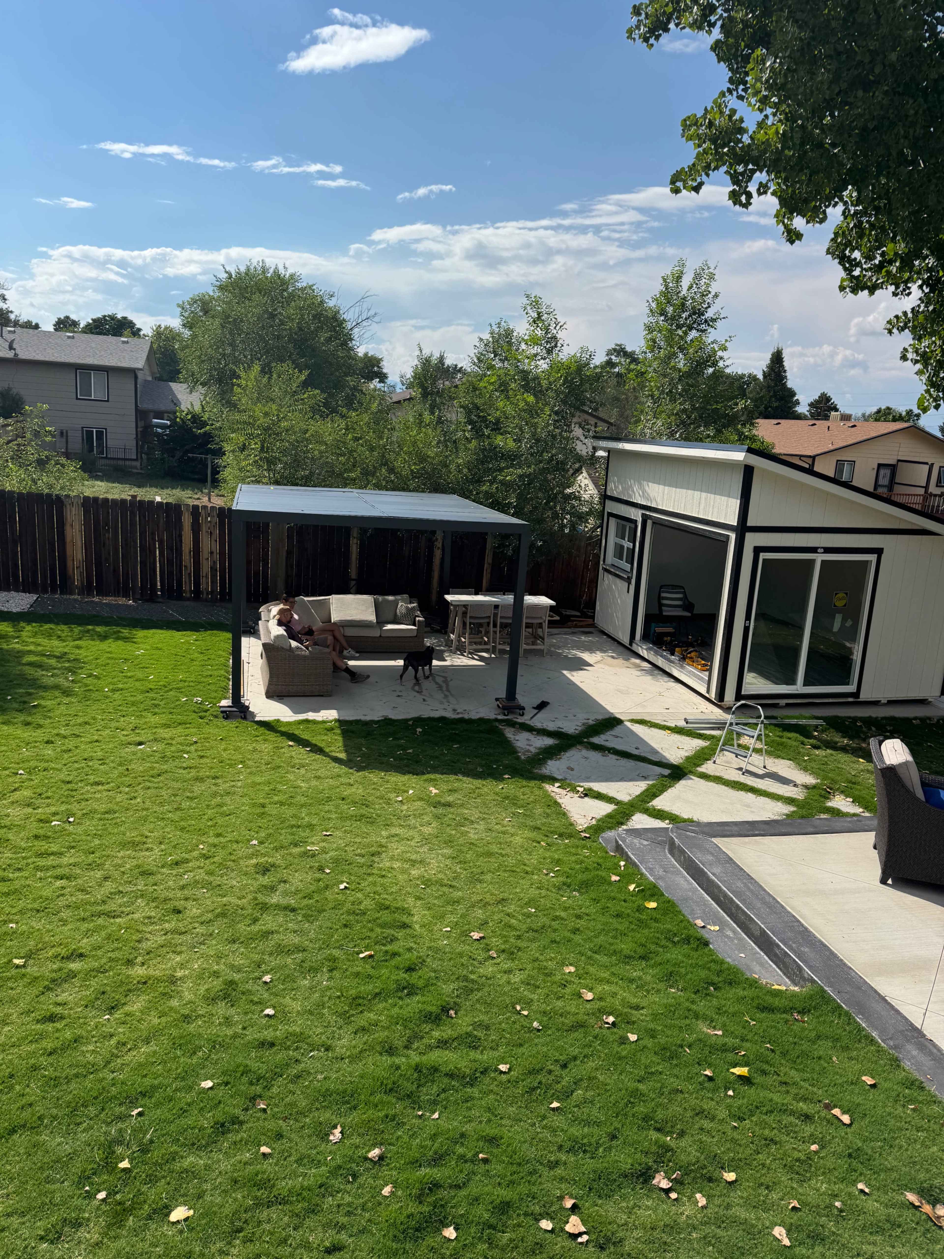 The image shows a backyard with a covered seating area, a lawn, and a separate shed or studio building in the background.