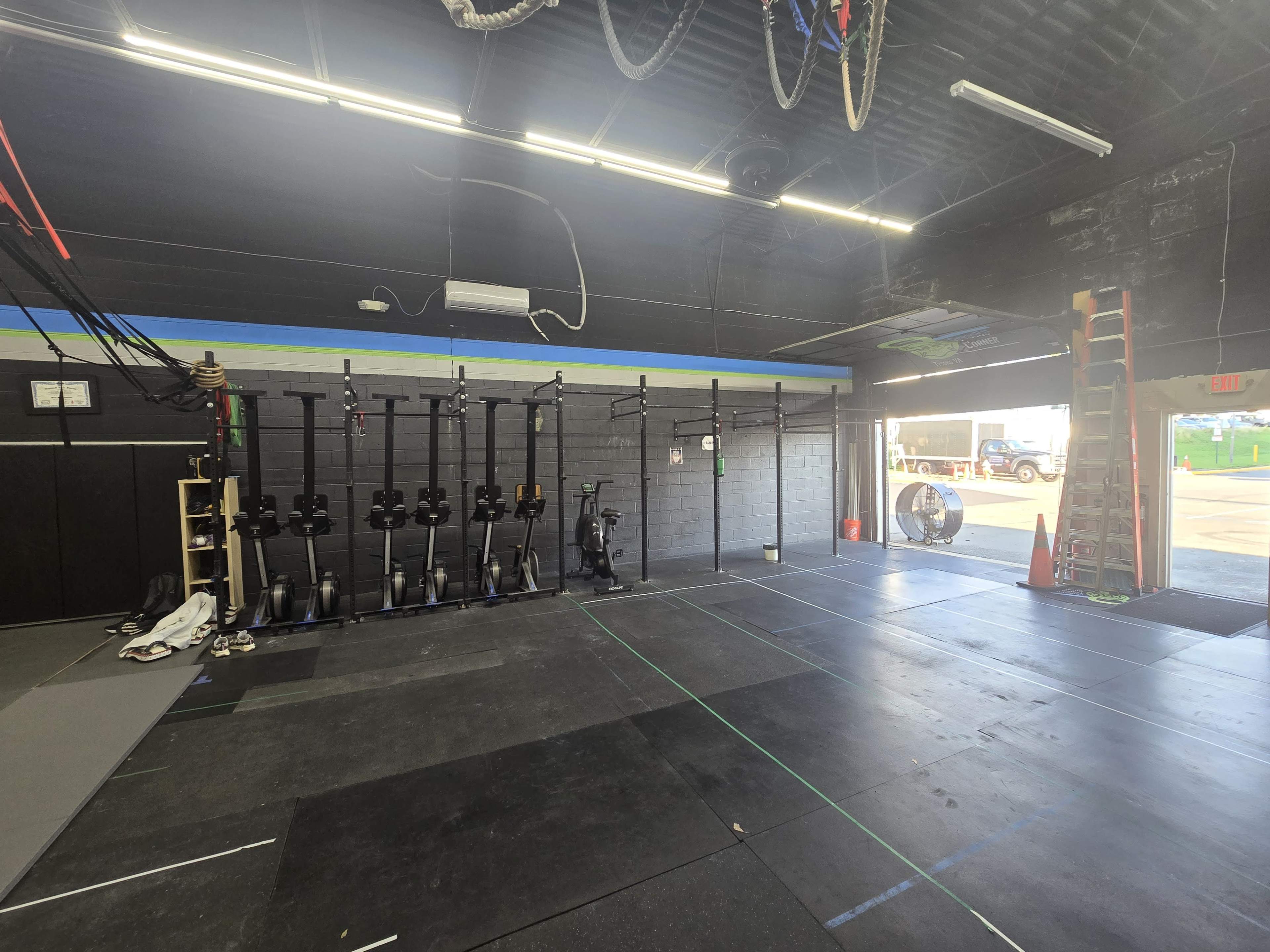 The image shows a gym interior with exercise equipment lined against the wall, including rowers, and a doorway leading outside.