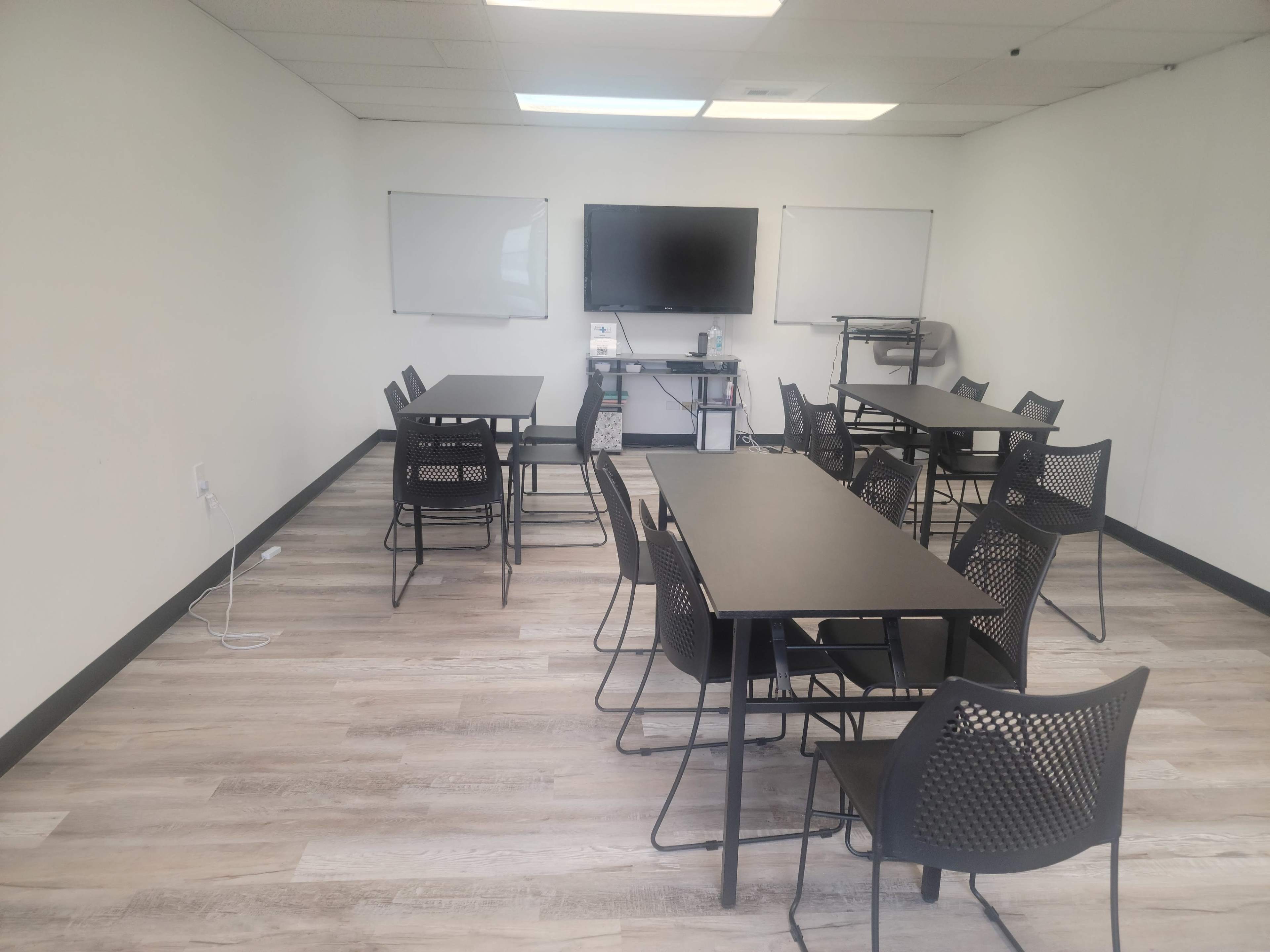 A conference room with multiple black tables and chairs, two whiteboards, and a large display screen on the wall.