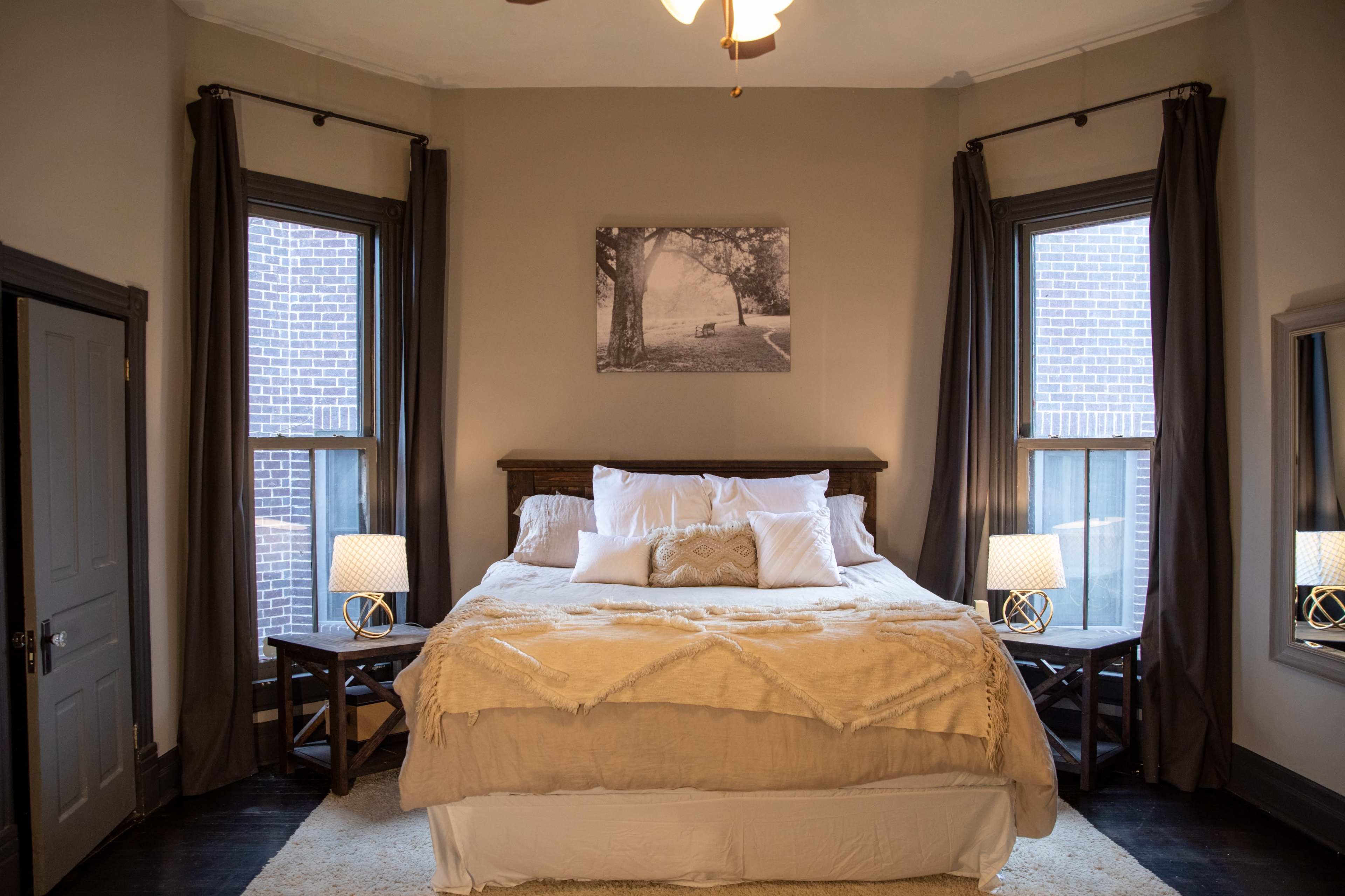 A well-decorated bedroom featuring a large bed with white linens, two nightstands with lamps, and windows adorned with dark curtains.