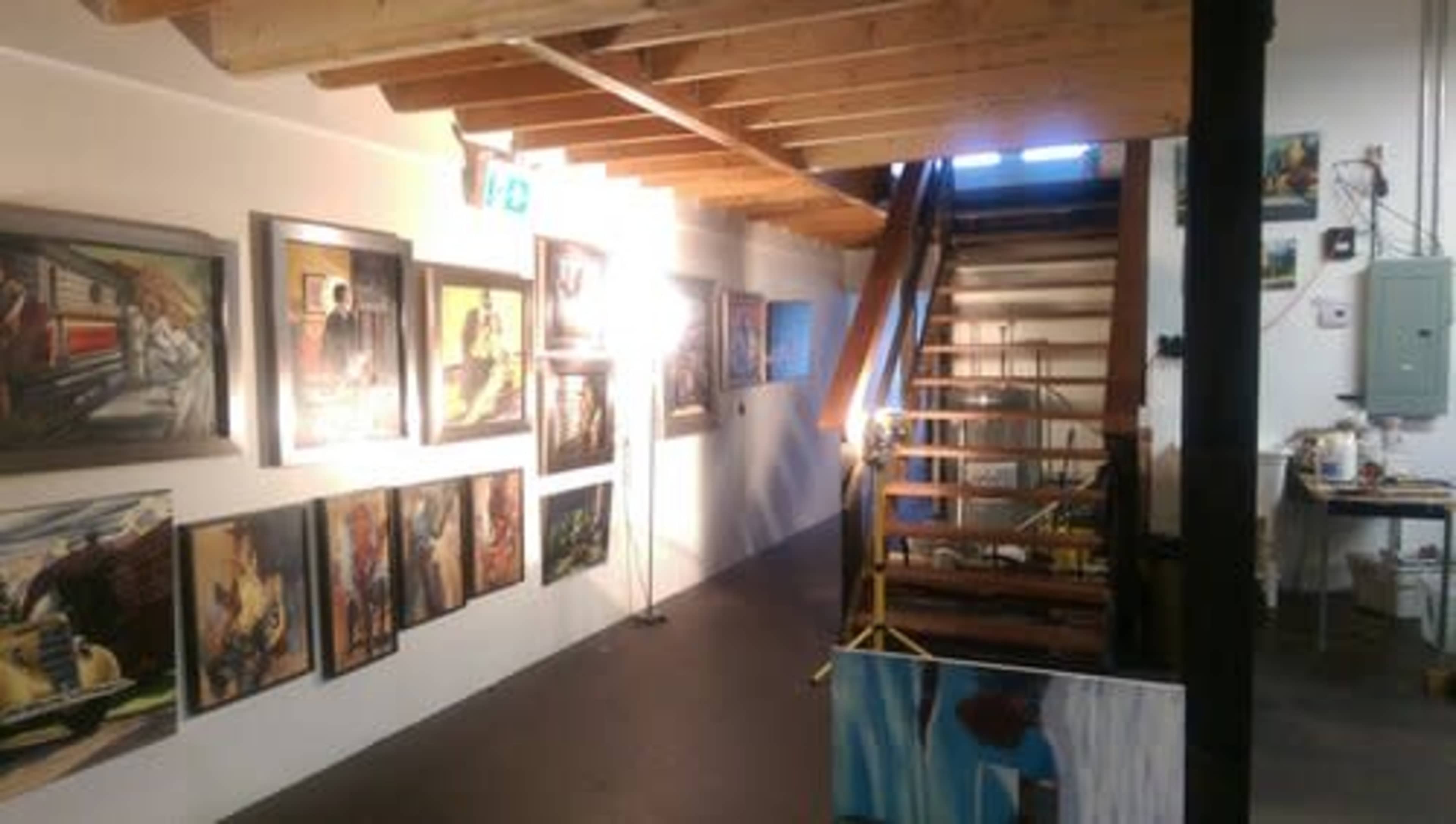 An art gallery hallway with framed paintings on the walls and a wooden staircase leading to a higher level.