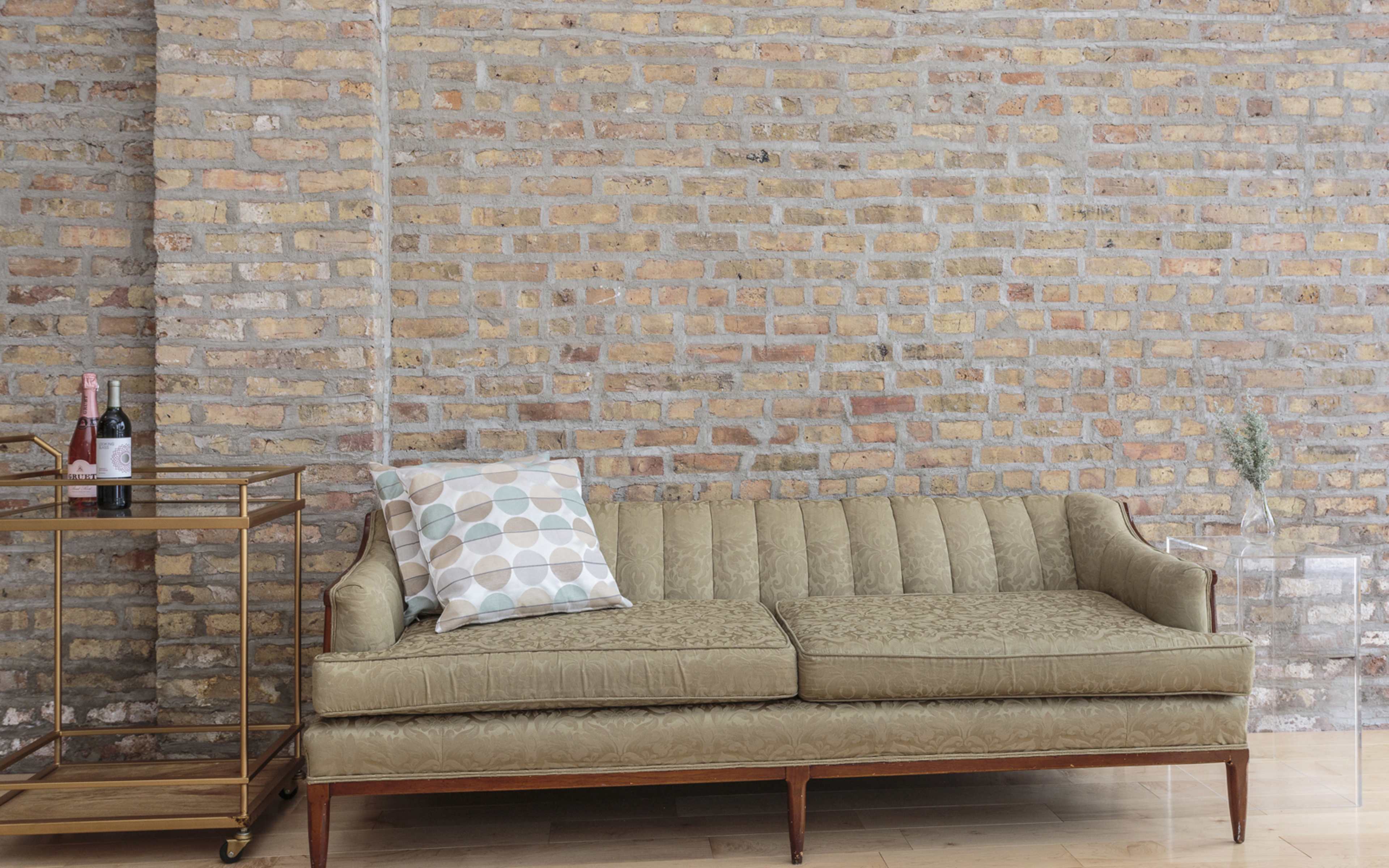 A green vintage sofa with a patterned pillow sits against a textured brick wall, beside a gold bar cart holding wine bottles.