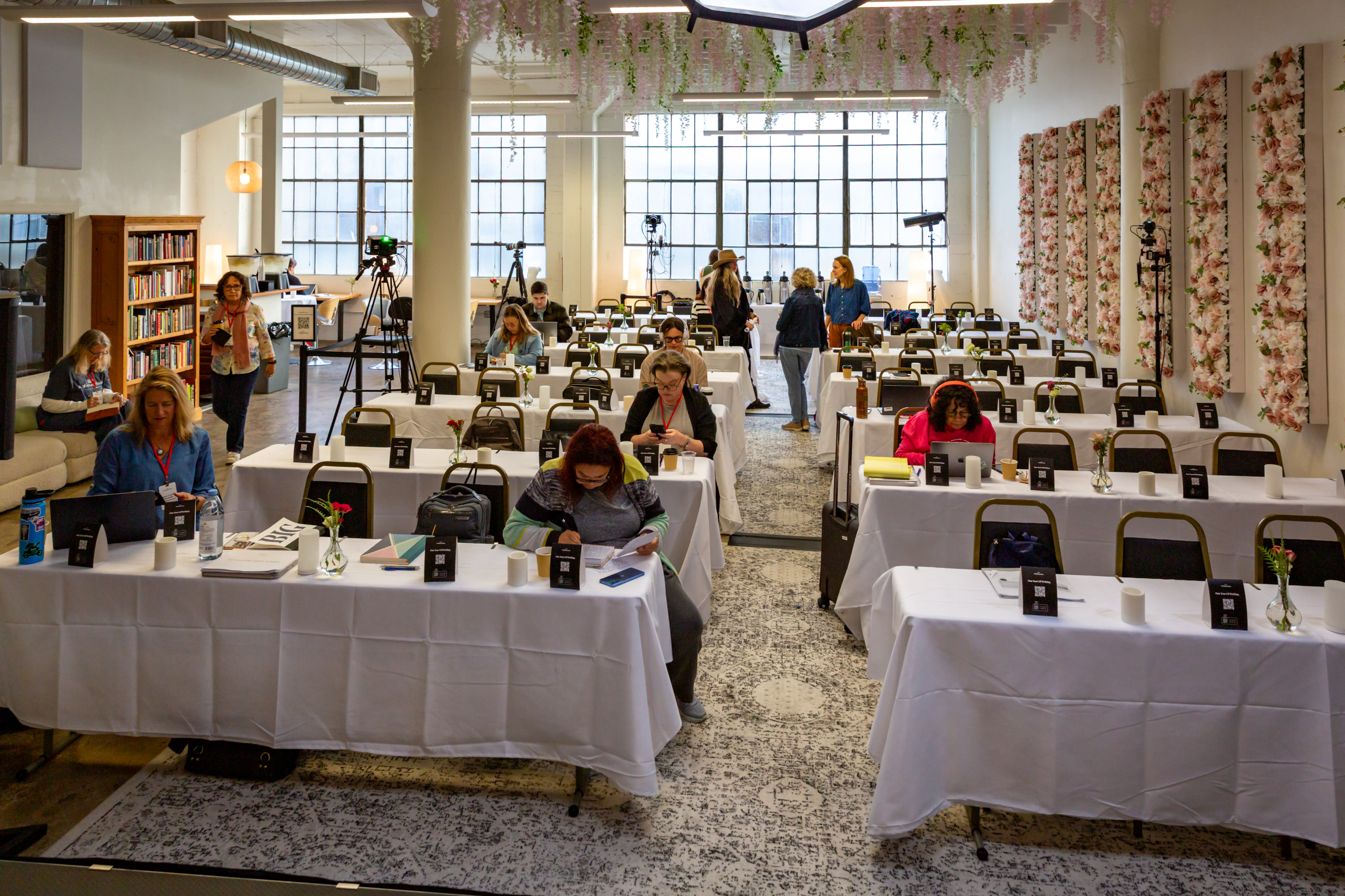 A spacious conference room is set up with rows of tables covered in white tablecloths, where attendees are actively engaged in discussions or working on laptops.
