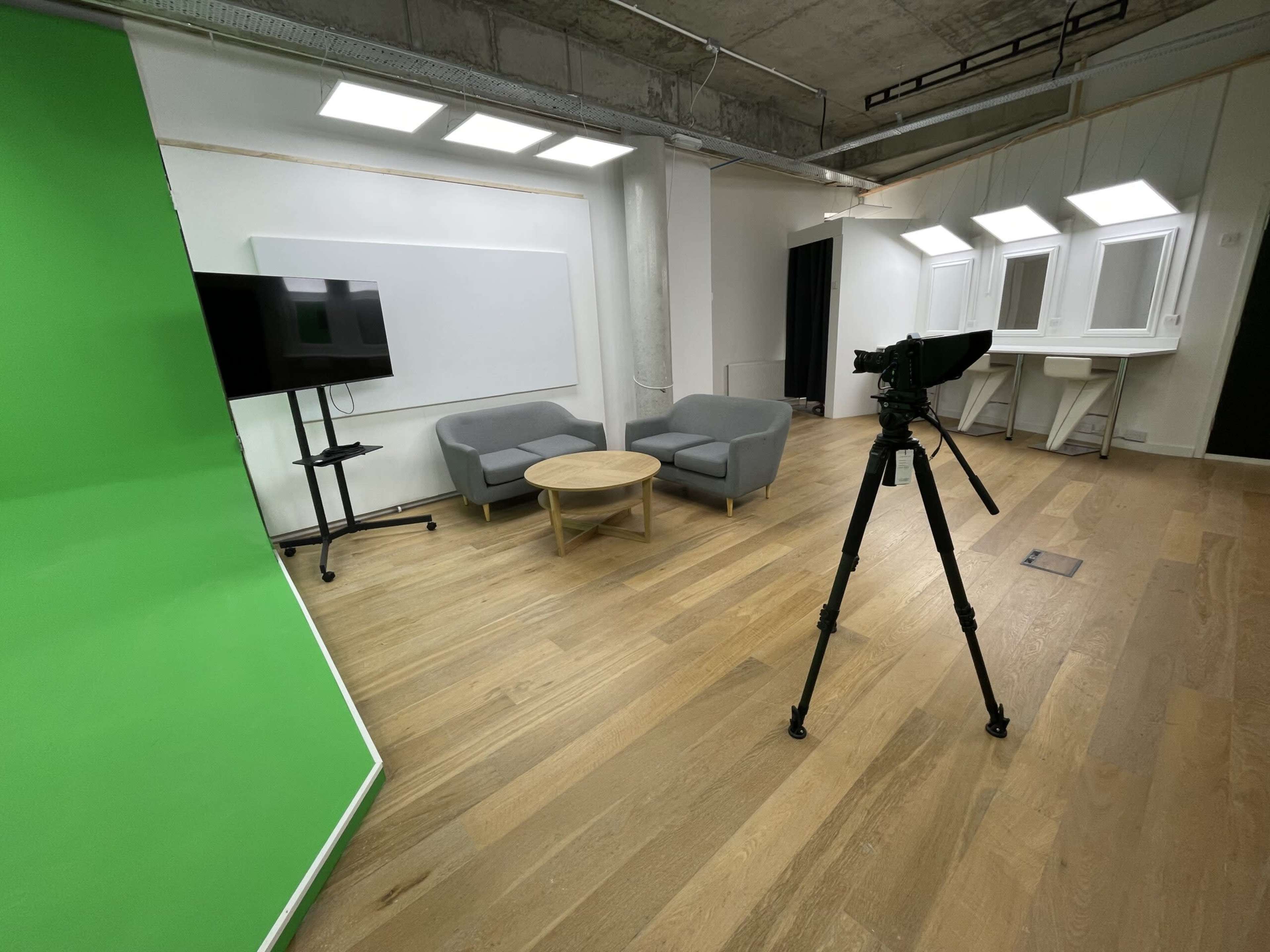 The image shows a studio setup with a green screen, a television, a round table, two gray chairs, and a camera on a tripod.