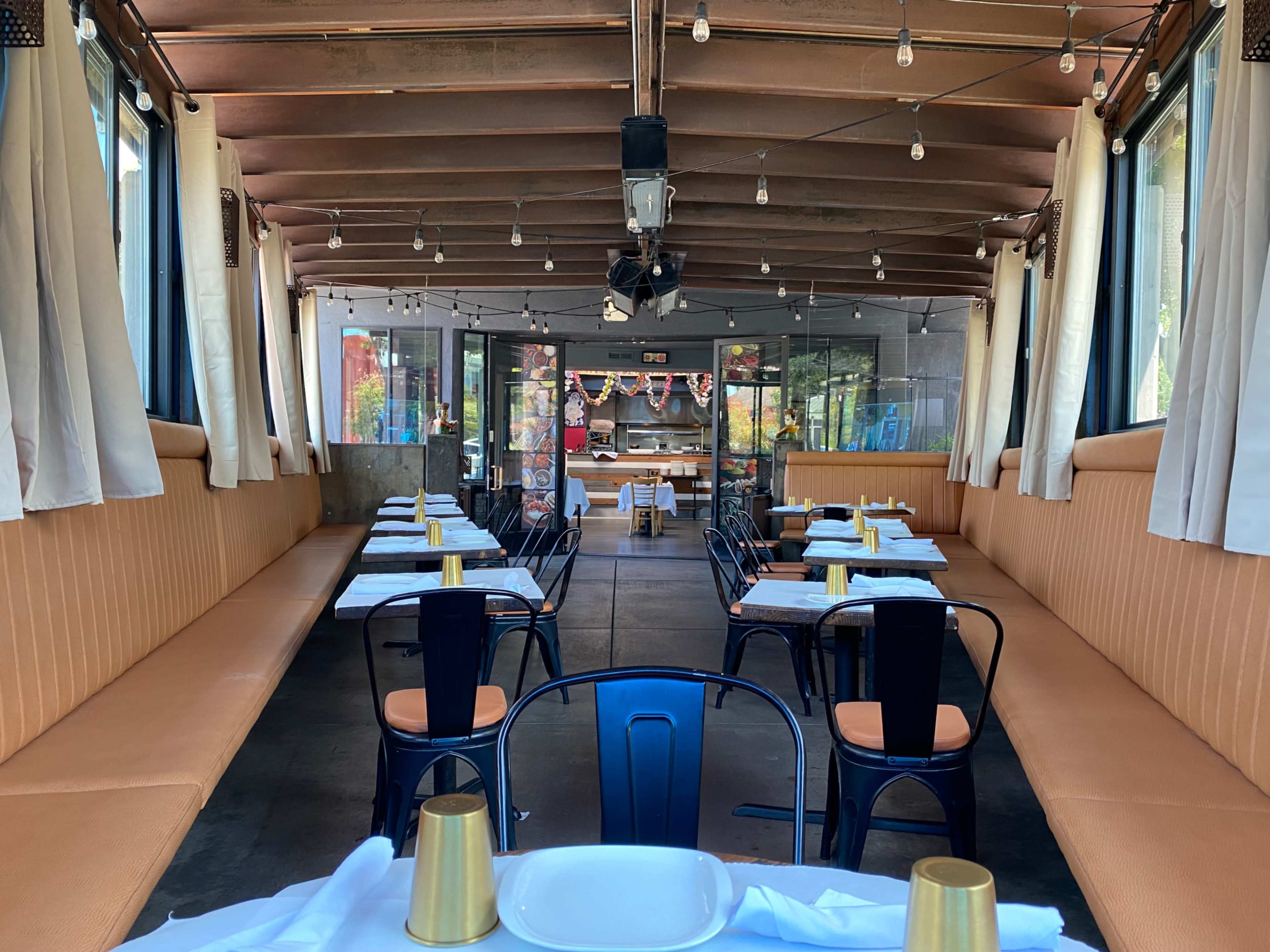 An empty restaurant interior featuring long booths, metal chairs, and a view of the kitchen area through large windows.