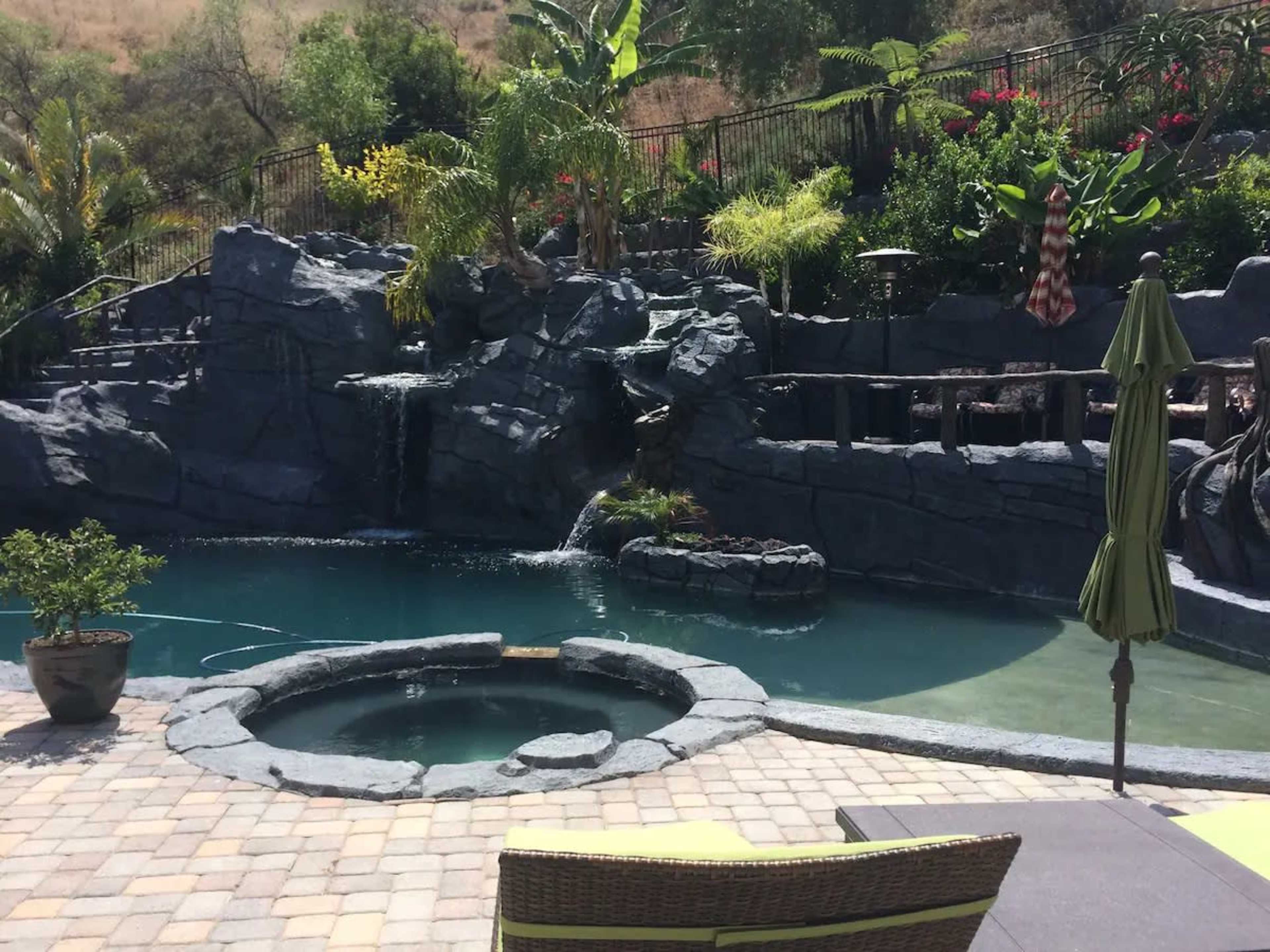 The image shows a landscaped pool area featuring a rock waterfall, a hot tub, and lush greenery.