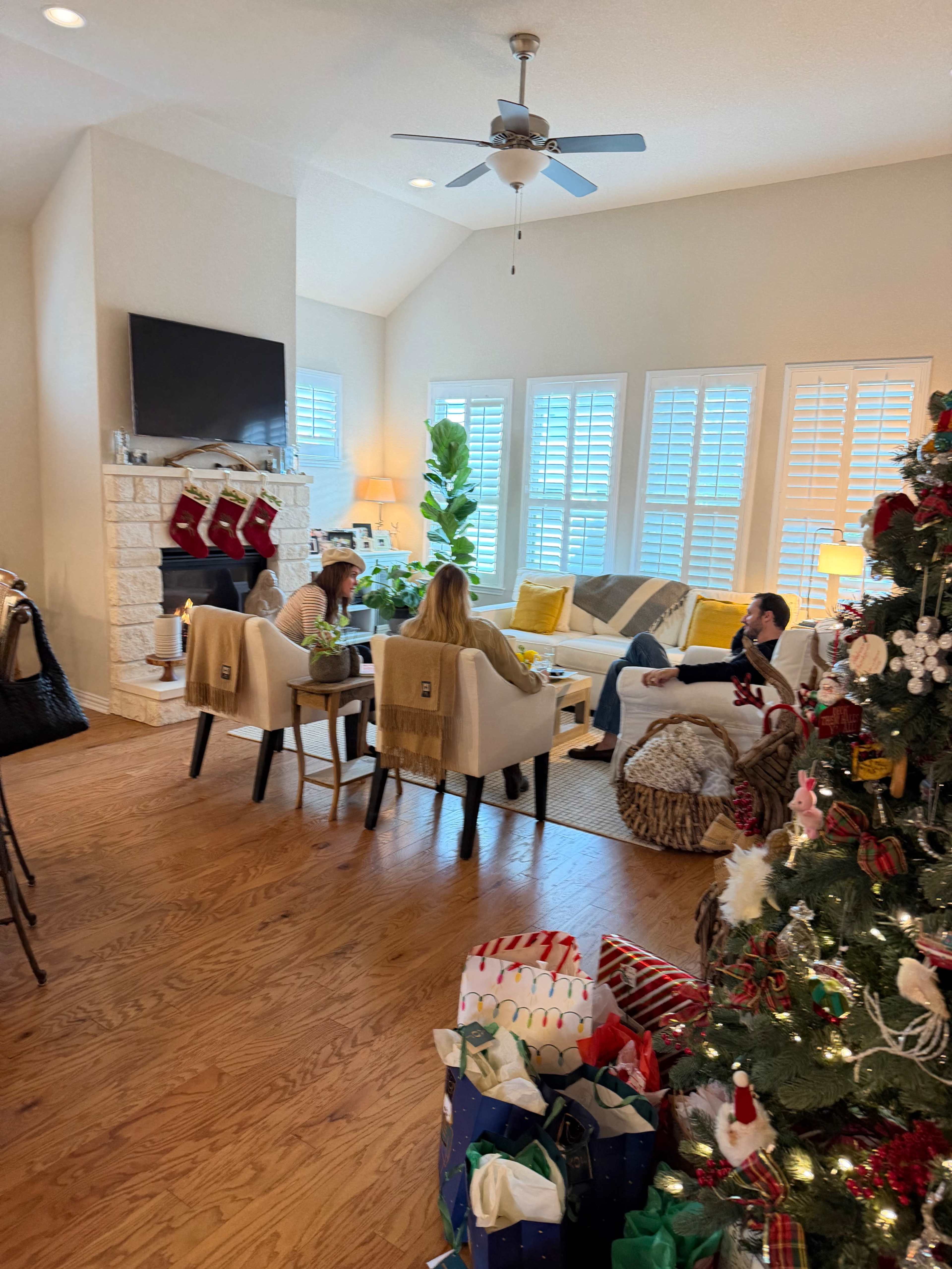 A cozy living room decorated for the holiday season features a tree adorned with gifts, a fireplace, and people sitting on a couch engaged in conversation.