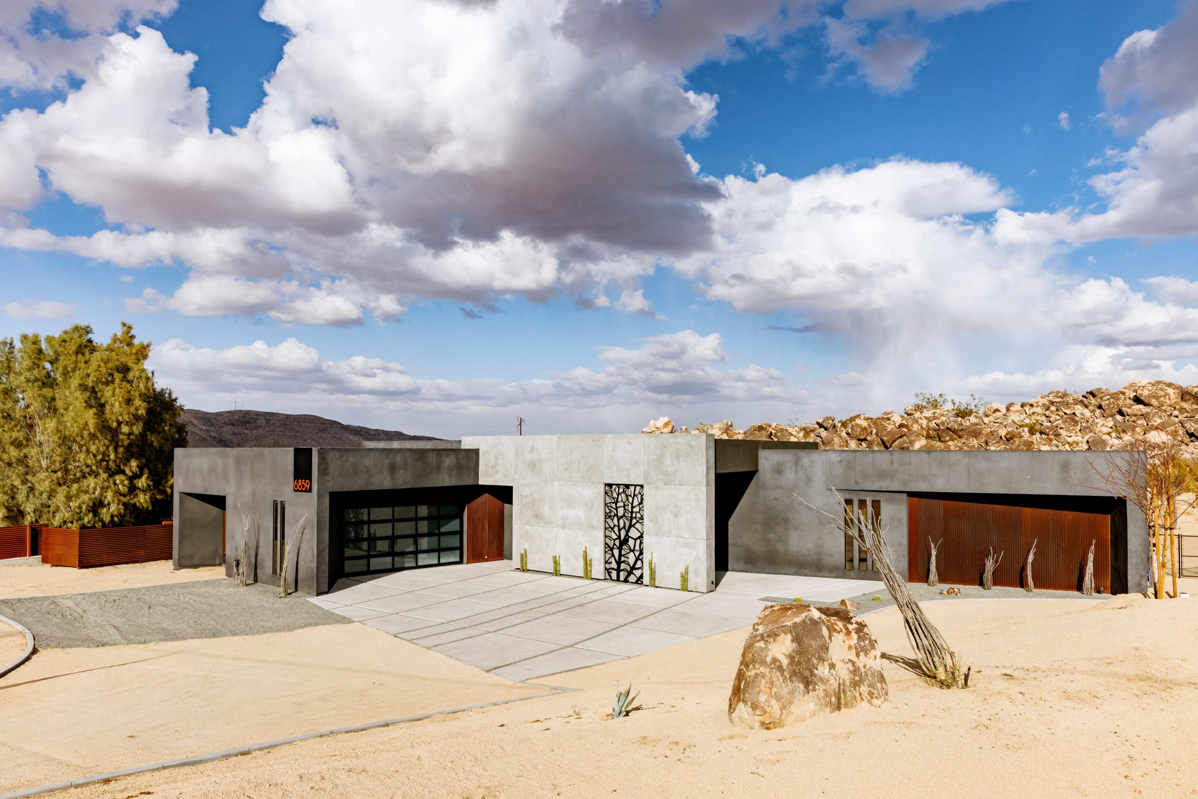 The image depicts a modern, angular house situated in a desert landscape with rocky formations and a partly cloudy sky.