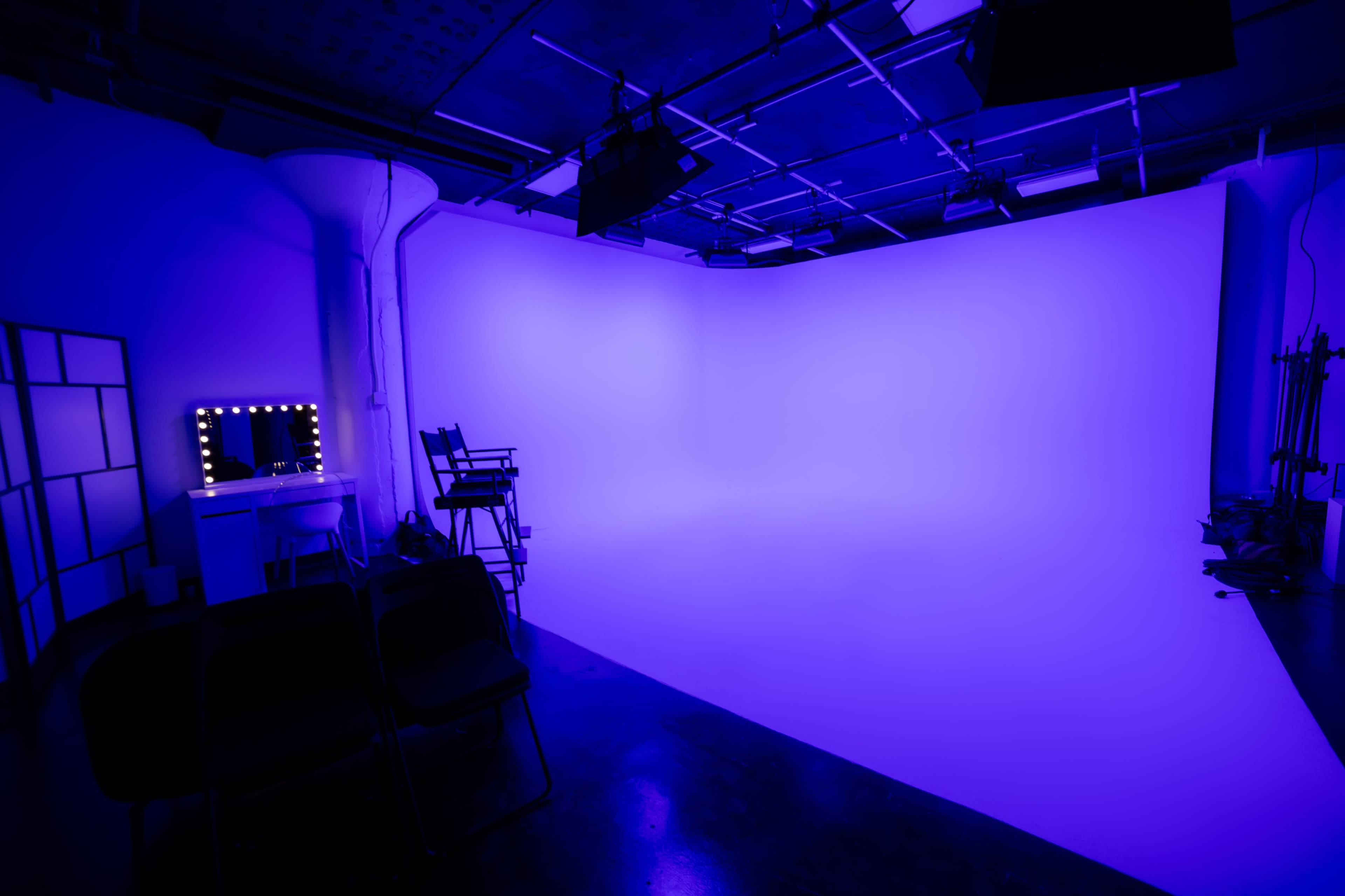 A studio space features a smooth, white backdrop illuminated with purple lighting, accompanied by a makeup mirror and a director's chair.