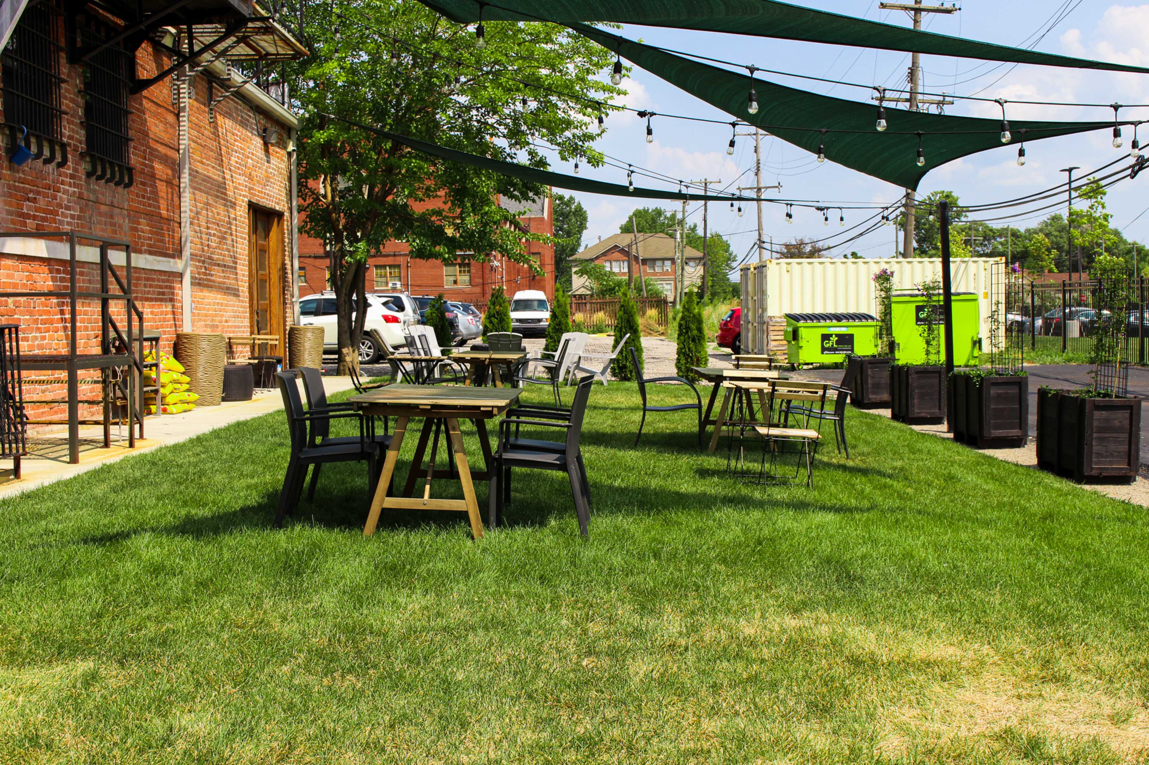 A grassy outdoor seating area with wooden tables and chairs, shaded by green canopies and surrounded by a brick building and parked vehicles.