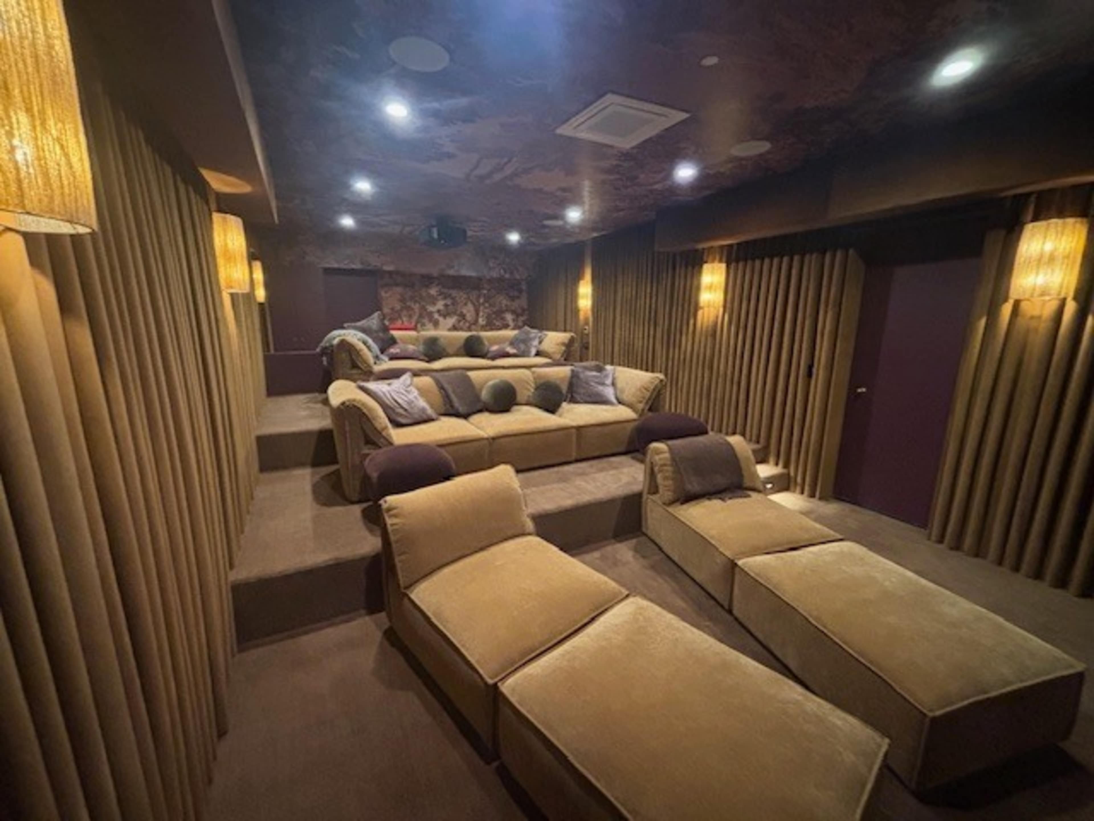 The image shows a home theater room with tiered seating featuring plush, upholstered sofas and chaise lounges, surrounded by textured walls and soft lighting.