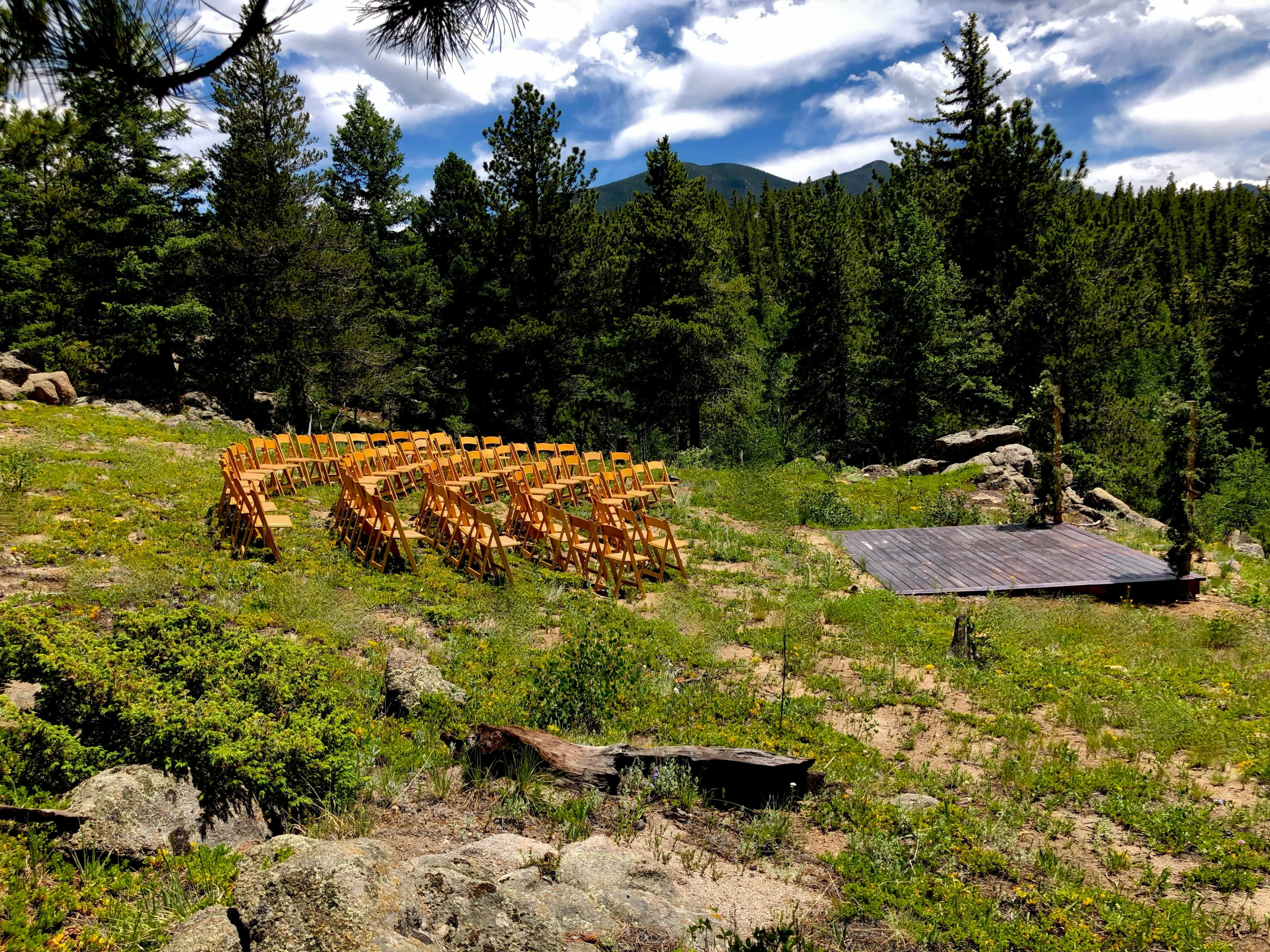 The image shows a clearing in a forest with rows of wooden chairs arranged for an event next to a small wooden stage.