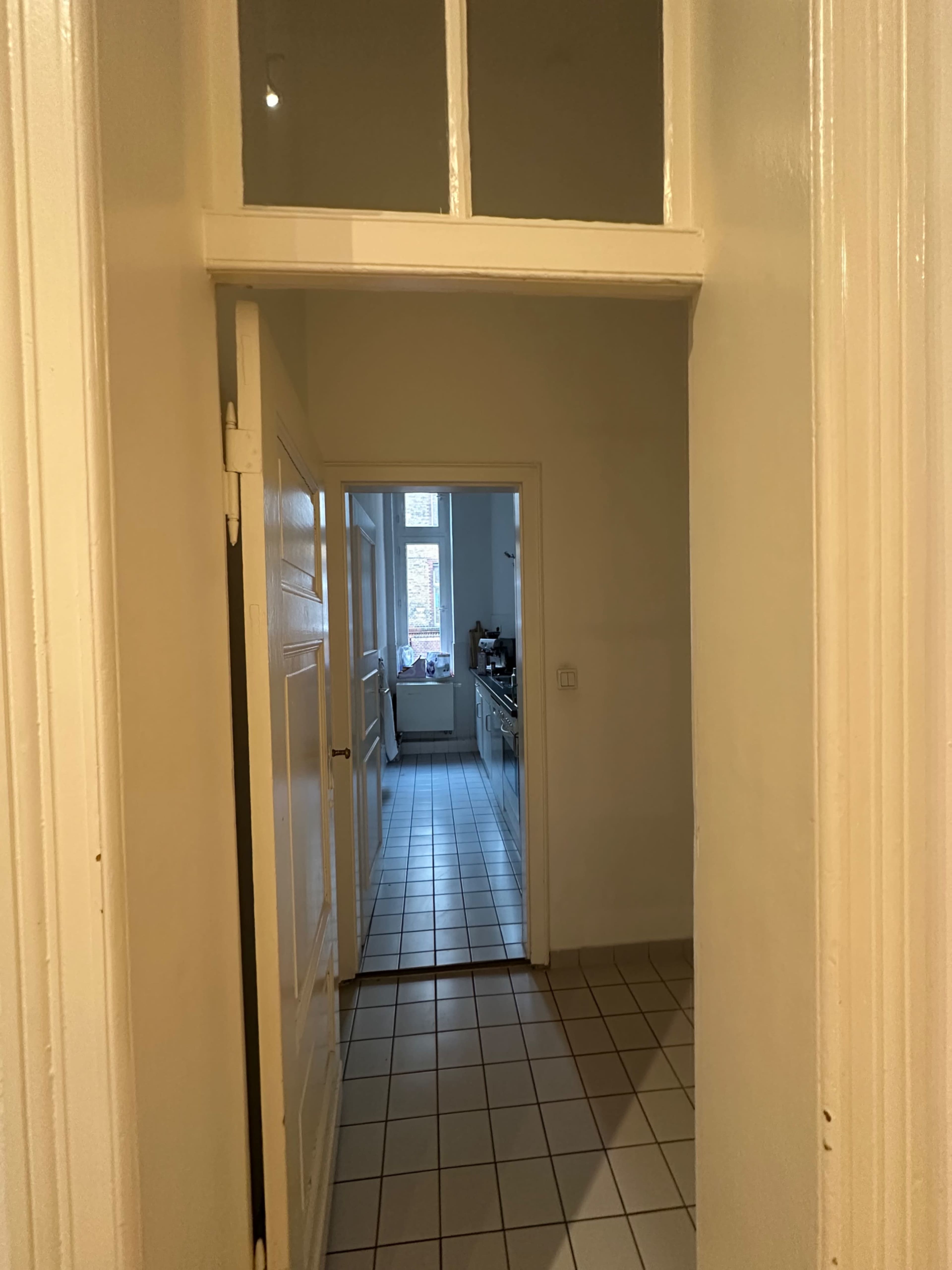 The image shows a hallway leading to a kitchen with tiled flooring and a doorway.
