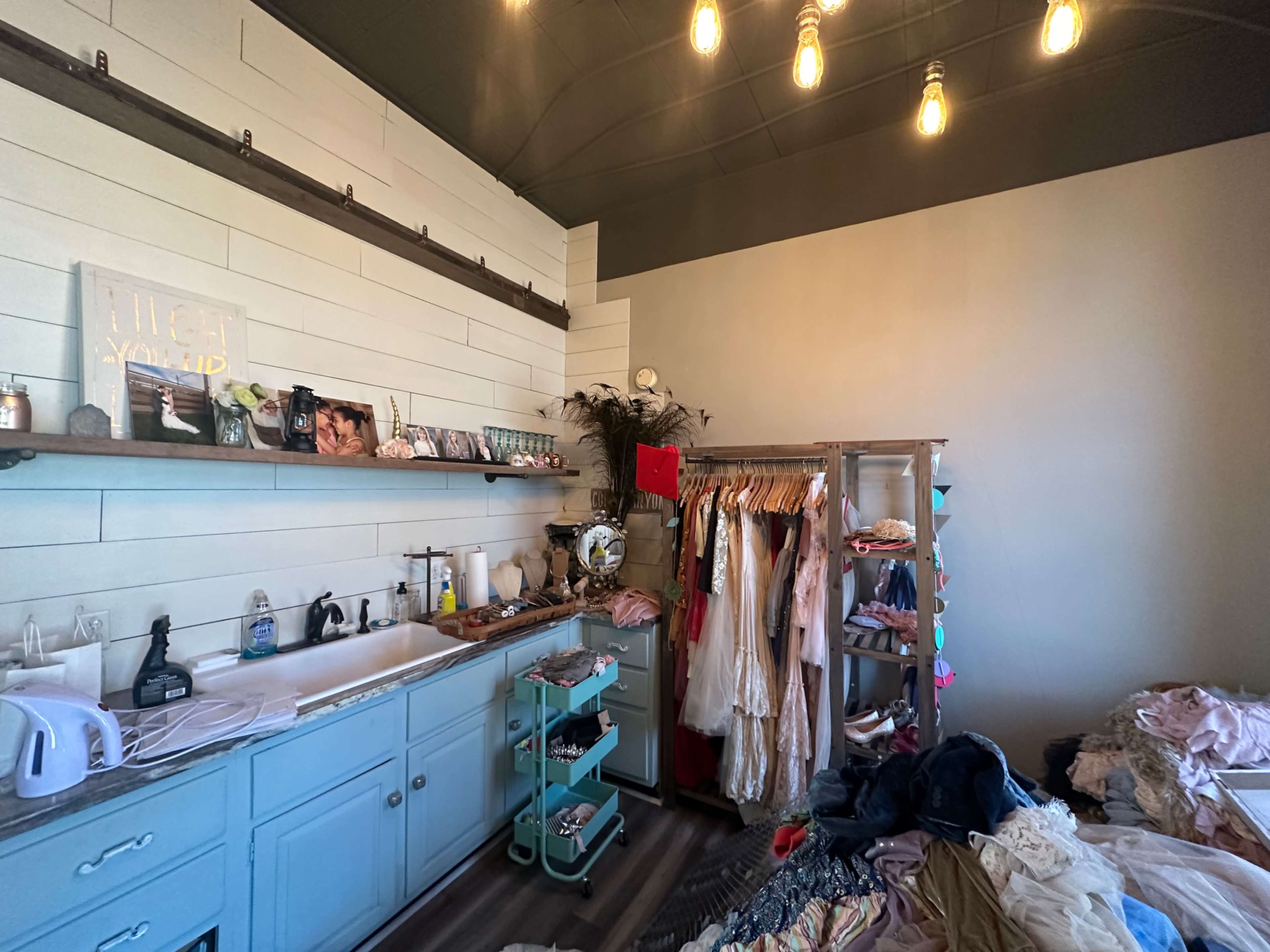 The image shows a room with a bed covered in clothes, a wooden clothing rack filled with garments, a light blue sink area, and shelves displaying various decorative items.