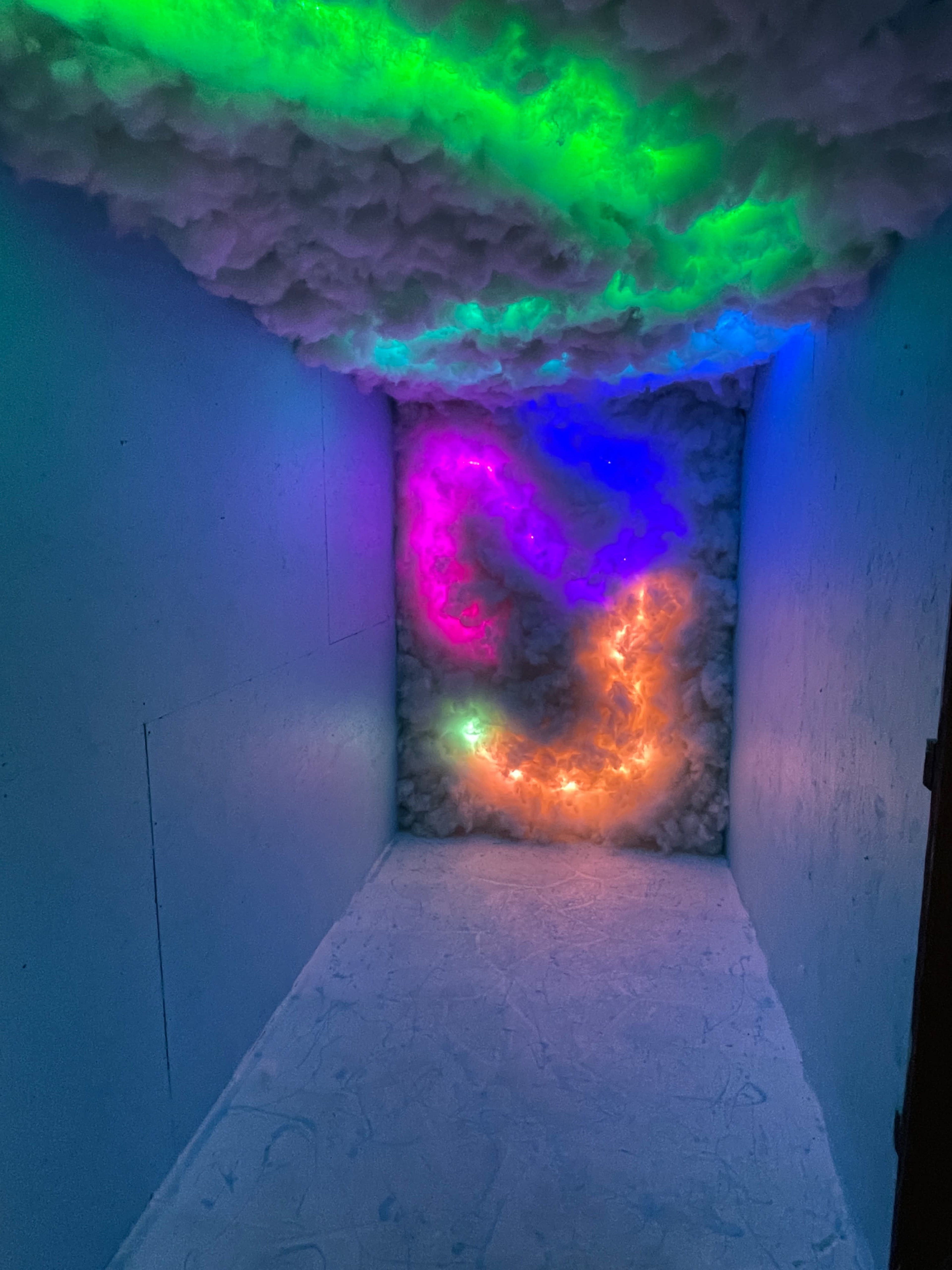 The image shows a dimly lit room with colorful, glowing patterns illuminated against a cloudy, textured wall.