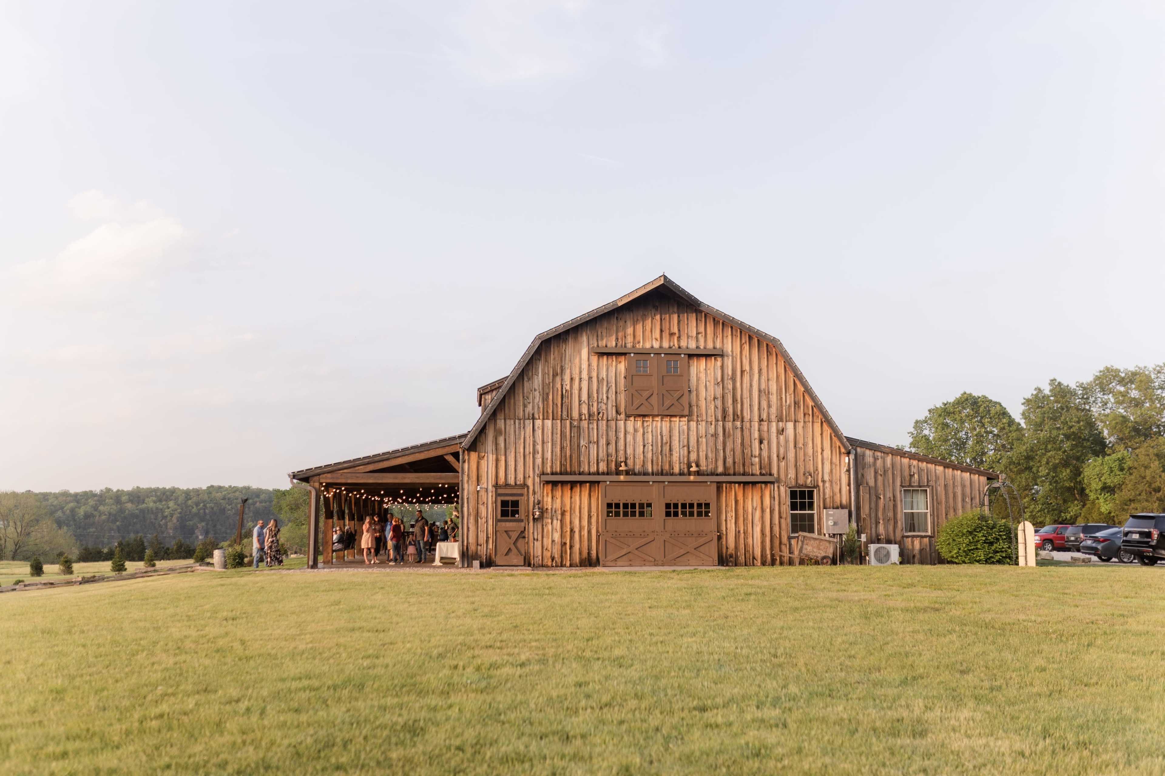 A rustic wooden barn stands in a grassy field under a clear sky, with twinkling lights visible inside and a gathering of people nearby.