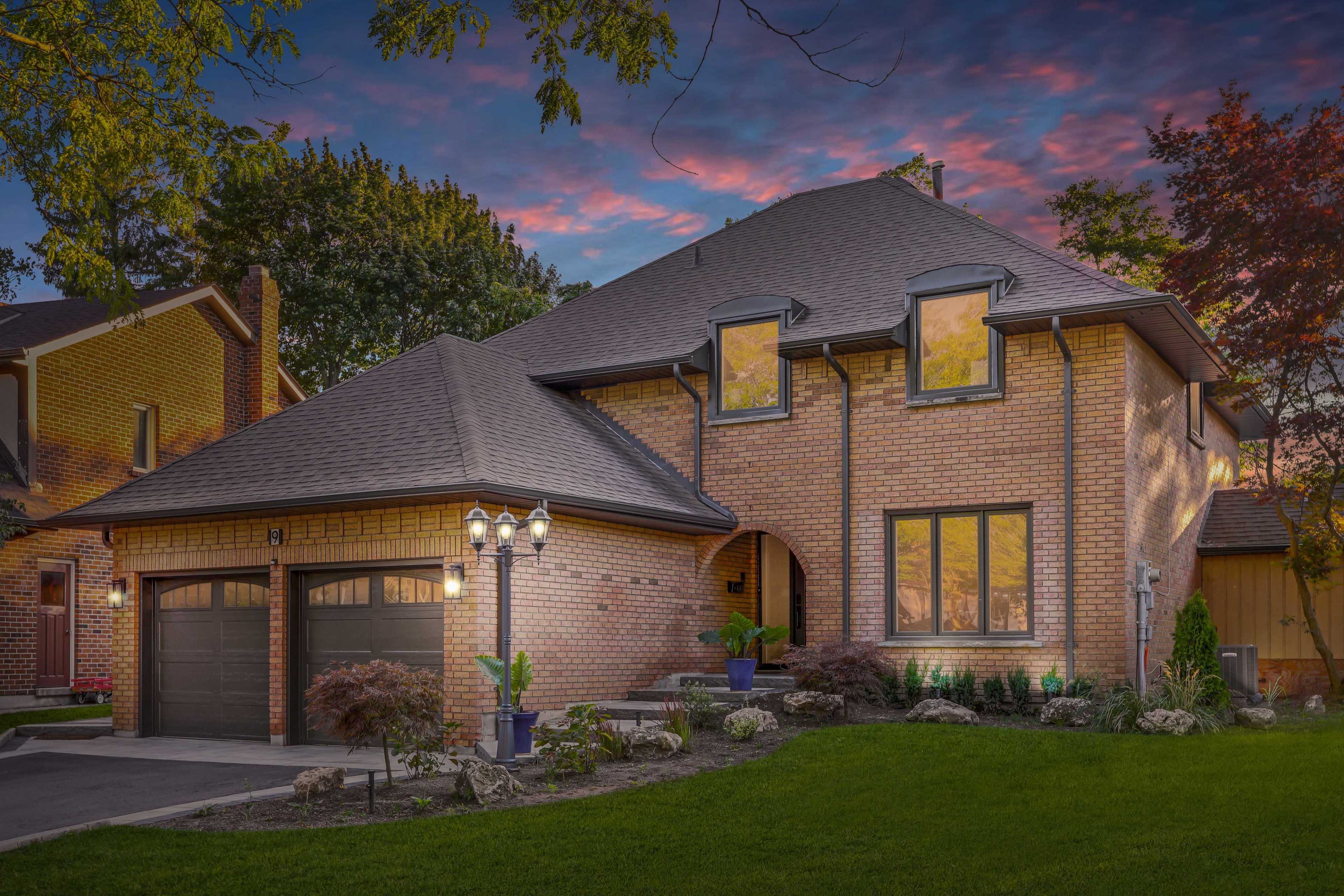 A two-story brick house with a curved entrance and large windows is surrounded by landscaped gardens under a colorful sky at dusk.